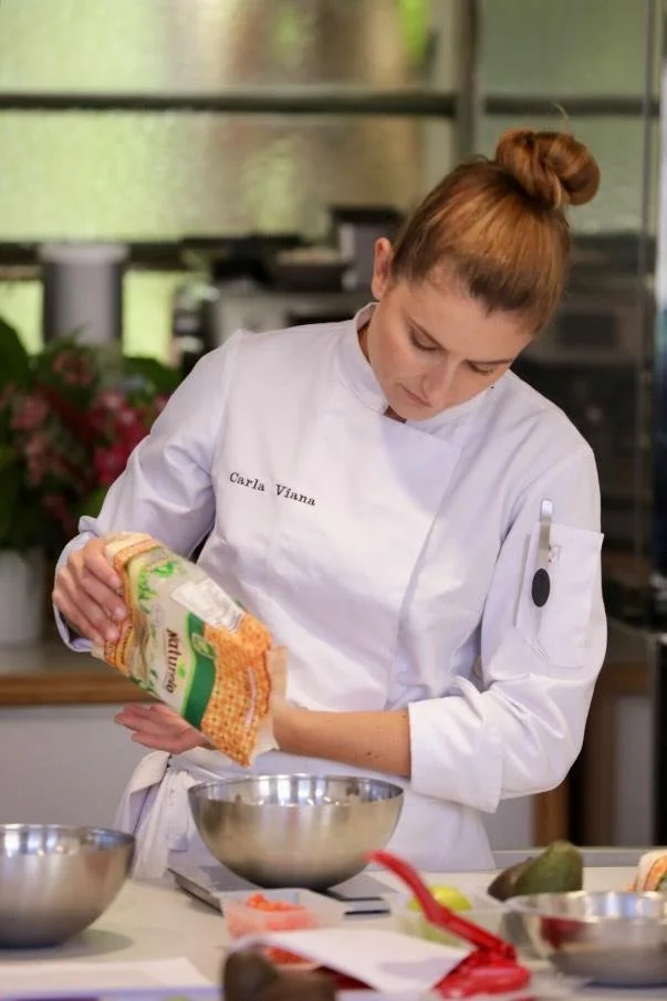 Carla Viana en veste de cuisine verse de la farine dans un bol en acier inoxydable dans une cuisine.