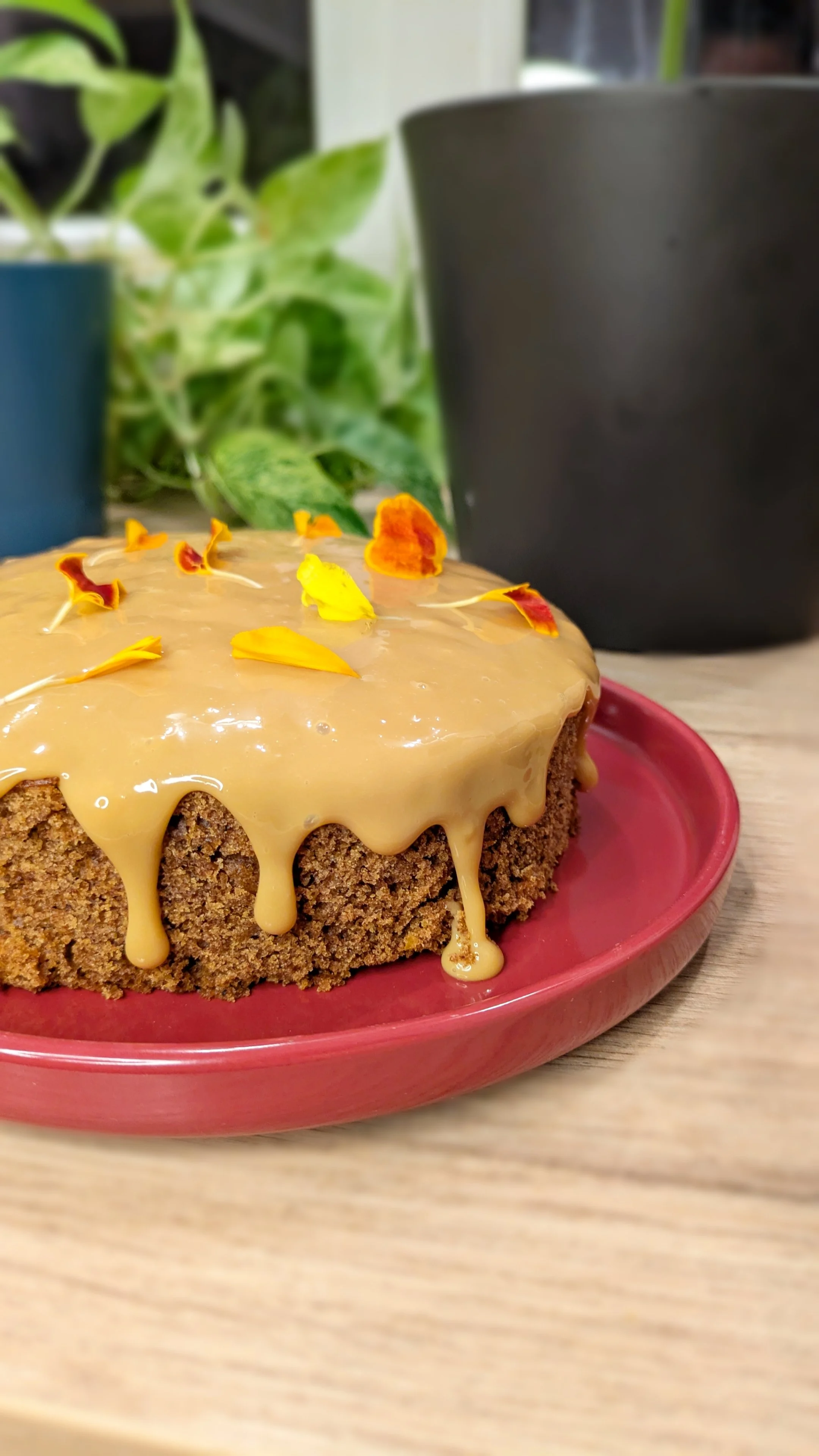 Gâteau avec une glaçure caramel, décoré de pétales de fleurs comestibles, dans un plat rouge, posé sur une table en bois.