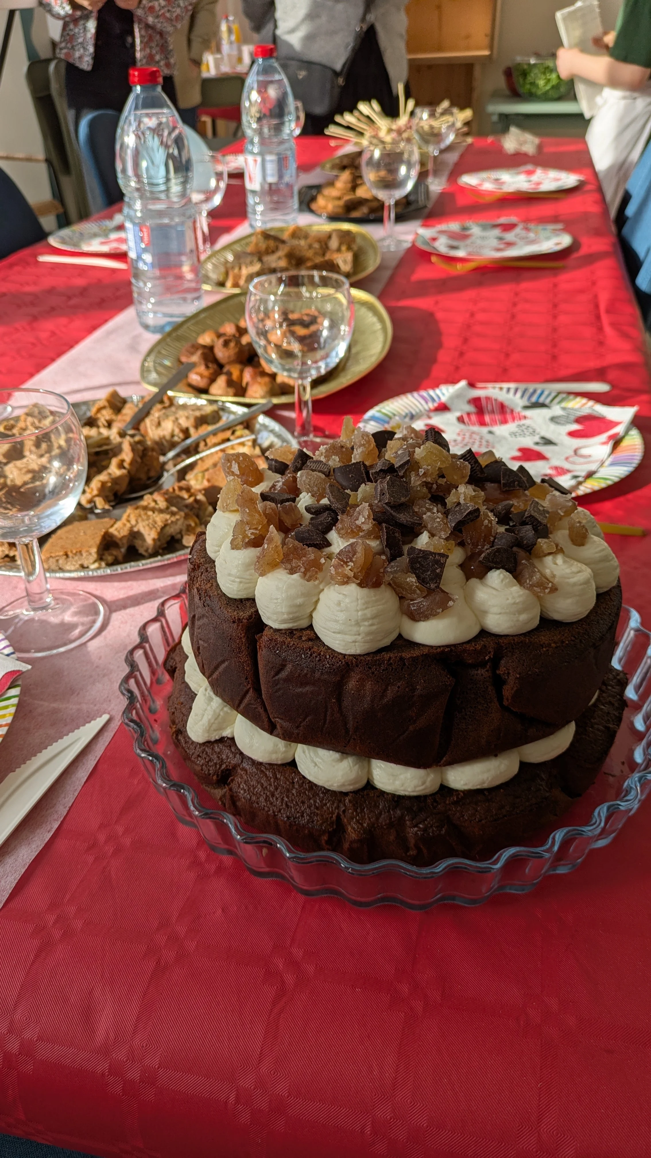 Un gâteau au chocolat décoré de crème fouettée et de morceaux de caramel, placé au centre d'une table avec d'autres plats et boissons, pour une fête ou un repas.