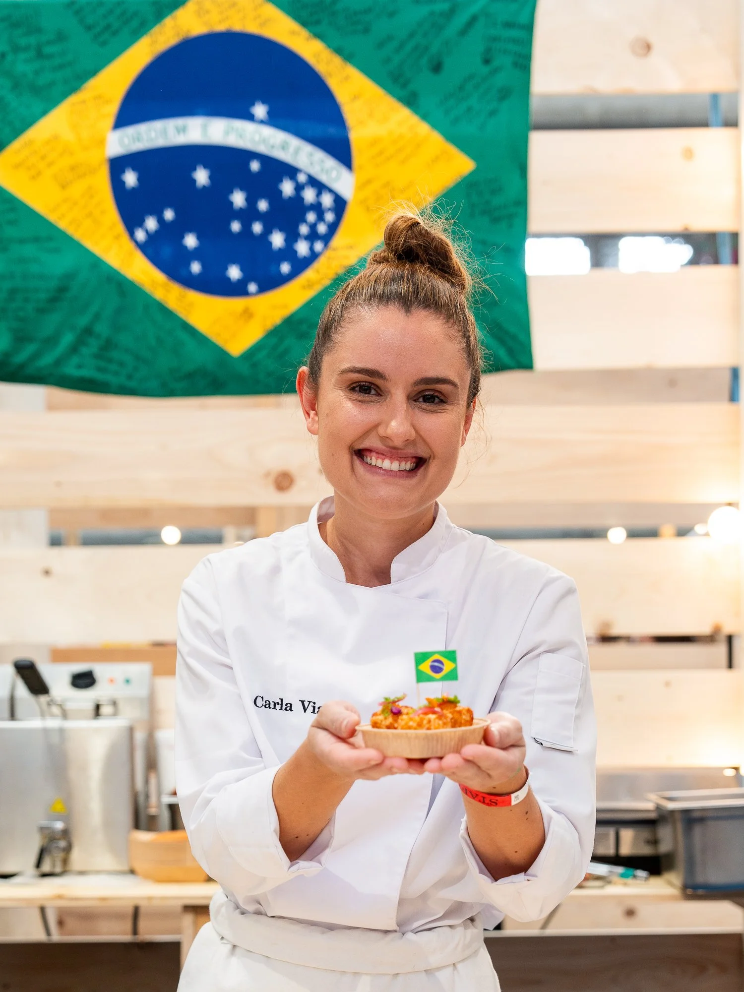 Une femme portant un uniforme blanc avec un badge marqué "Carla V" sourit et tient un petit plat avec de la nourriture, devant un drapeau du Brésil. 