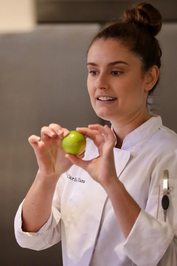Carla Viana en uniforme de chef cuisinier tient un citron vert dans ses mains, en train de l'examiner.