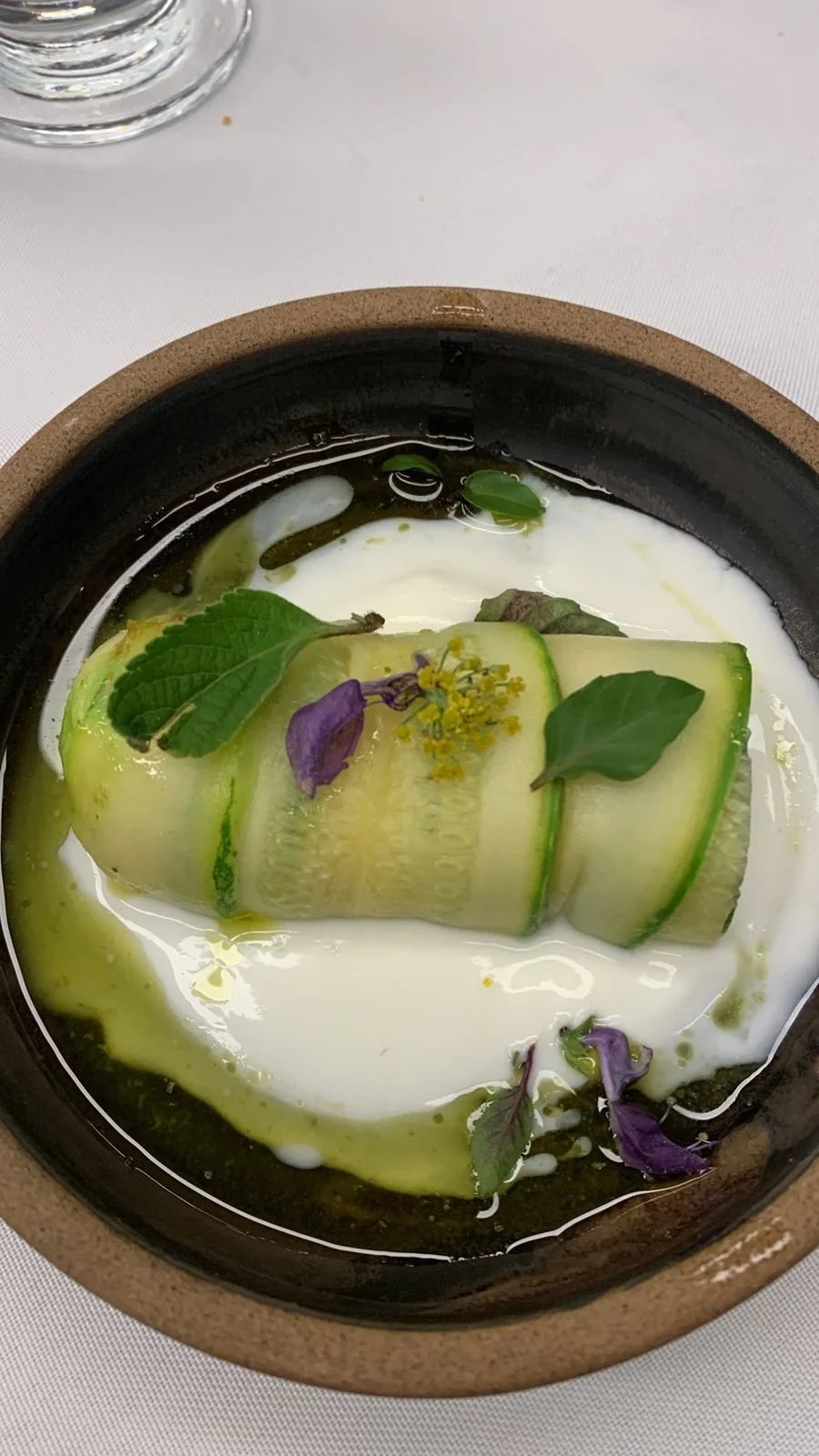 Un plat gastronomique avec des tranches de courgette, des feuilles de menthe, des fleurs violettes et jaunes, dans un bol en terre cuite avec une sauce blanche, décoré de manière élégante.