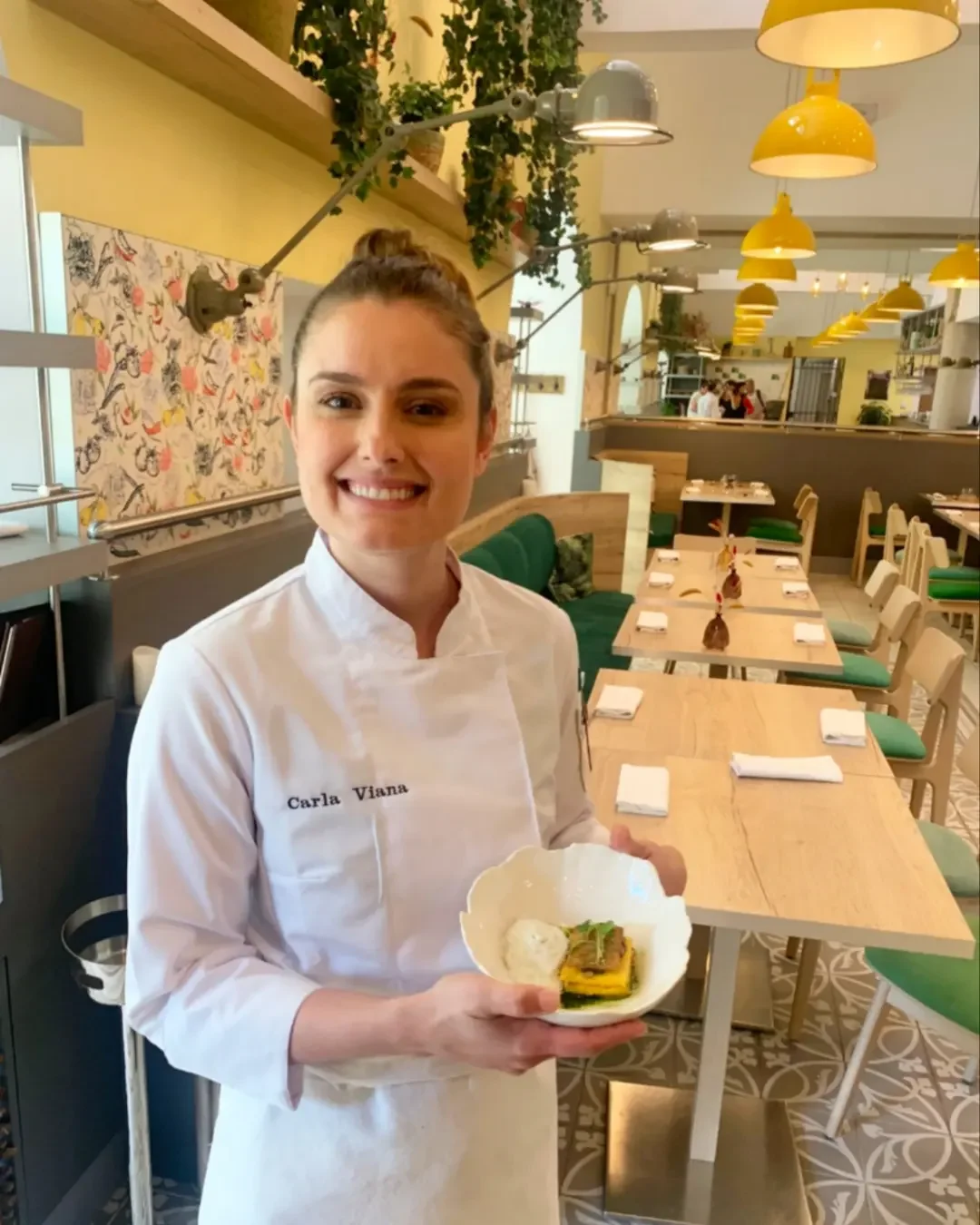 Une femme souriante, portant un uniforme de chef avec le nom Carla Viana, tient une assiette contenant un plat de cuisine dans un restaurant moderne avec une décoration colorée et lumineuse.