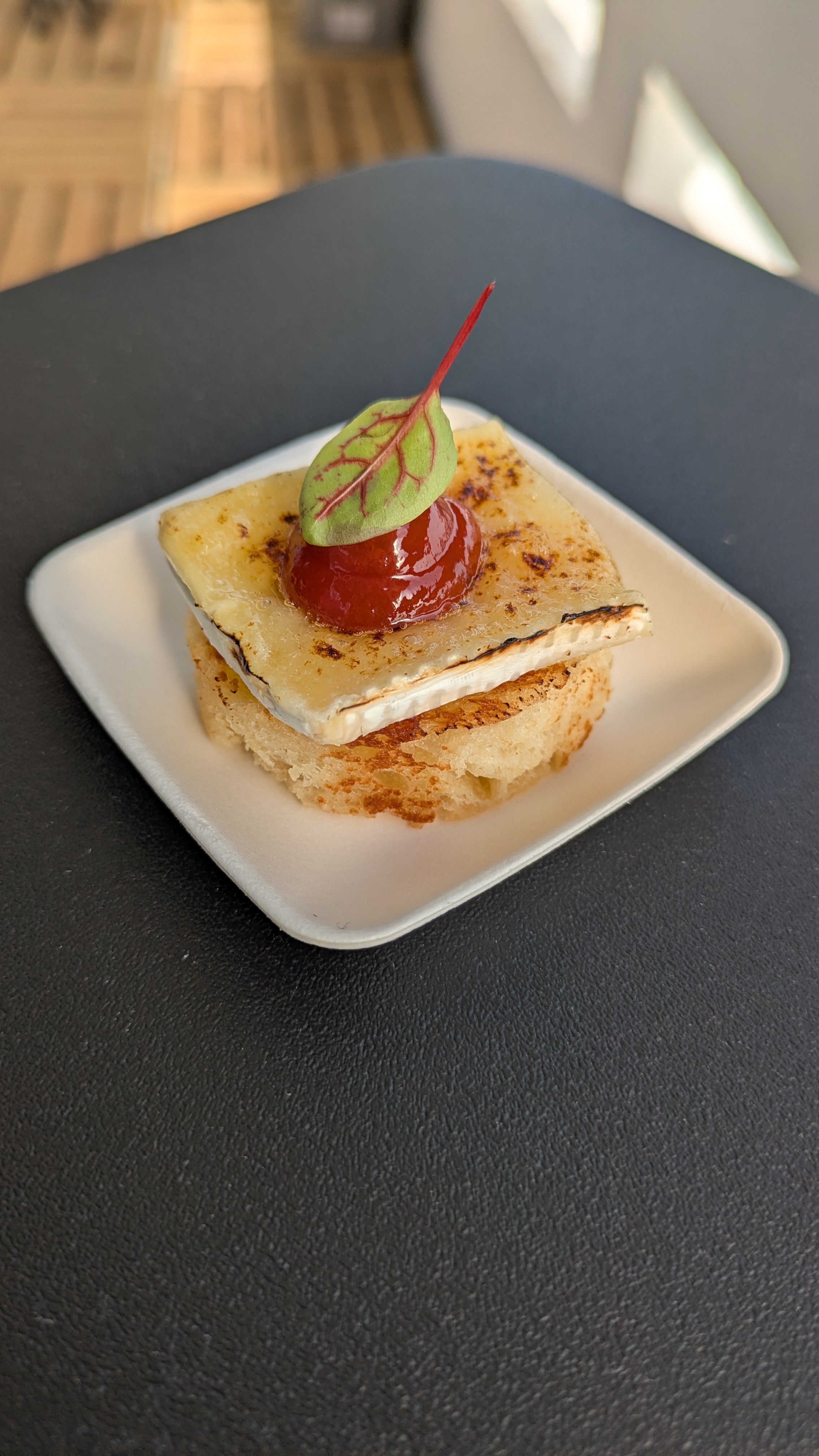 Canapé avec fromage, un cercle de confiture de framboise, une petite feuille de salade, présenté sur une assiette blanche, posé sur une surface noire.