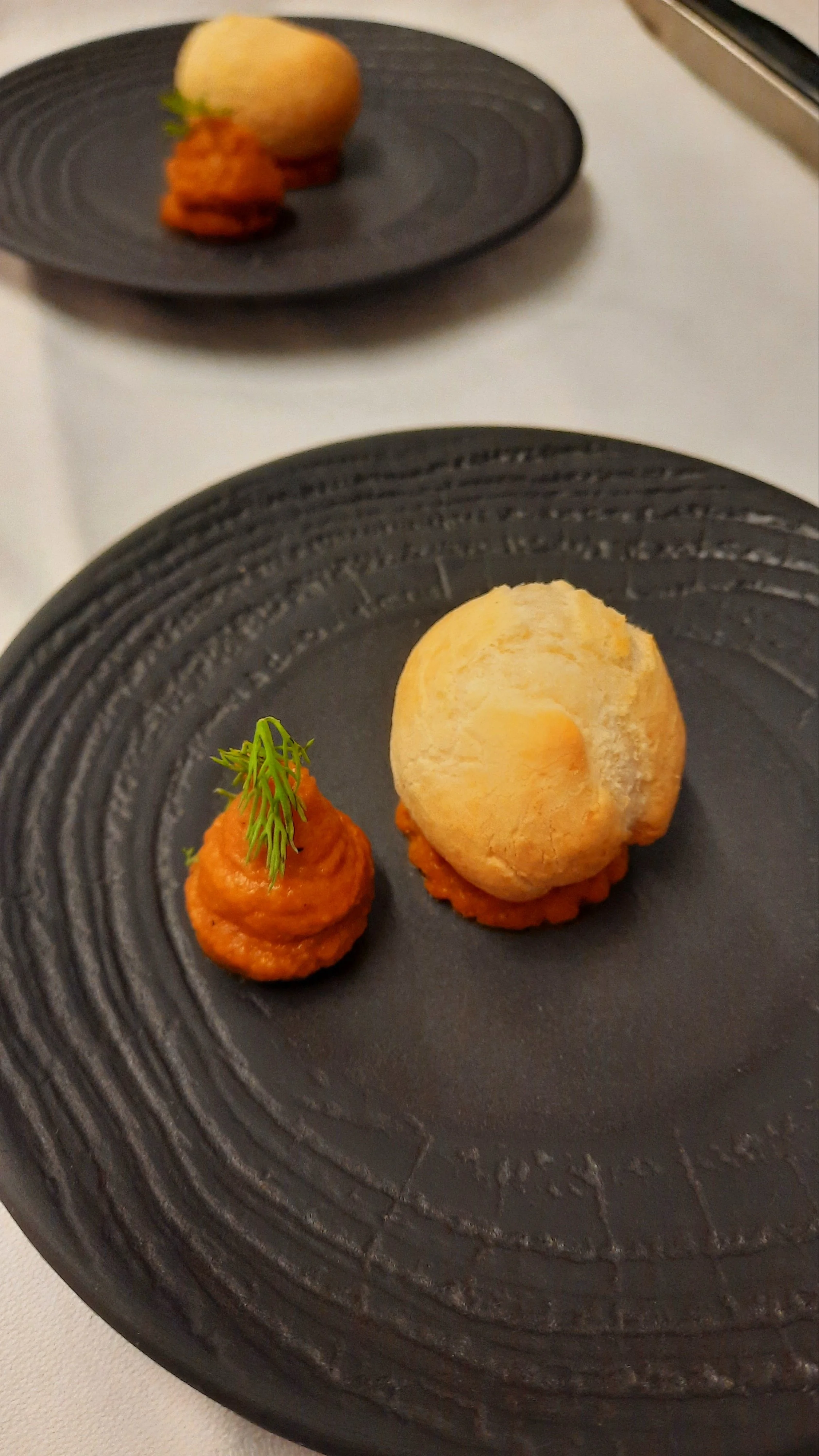 Deux petits plats présentés sur une assiette noire, un avec une boule de pain et l'autre avec une purée de légumes, décorés d'une petite branche de verdure.