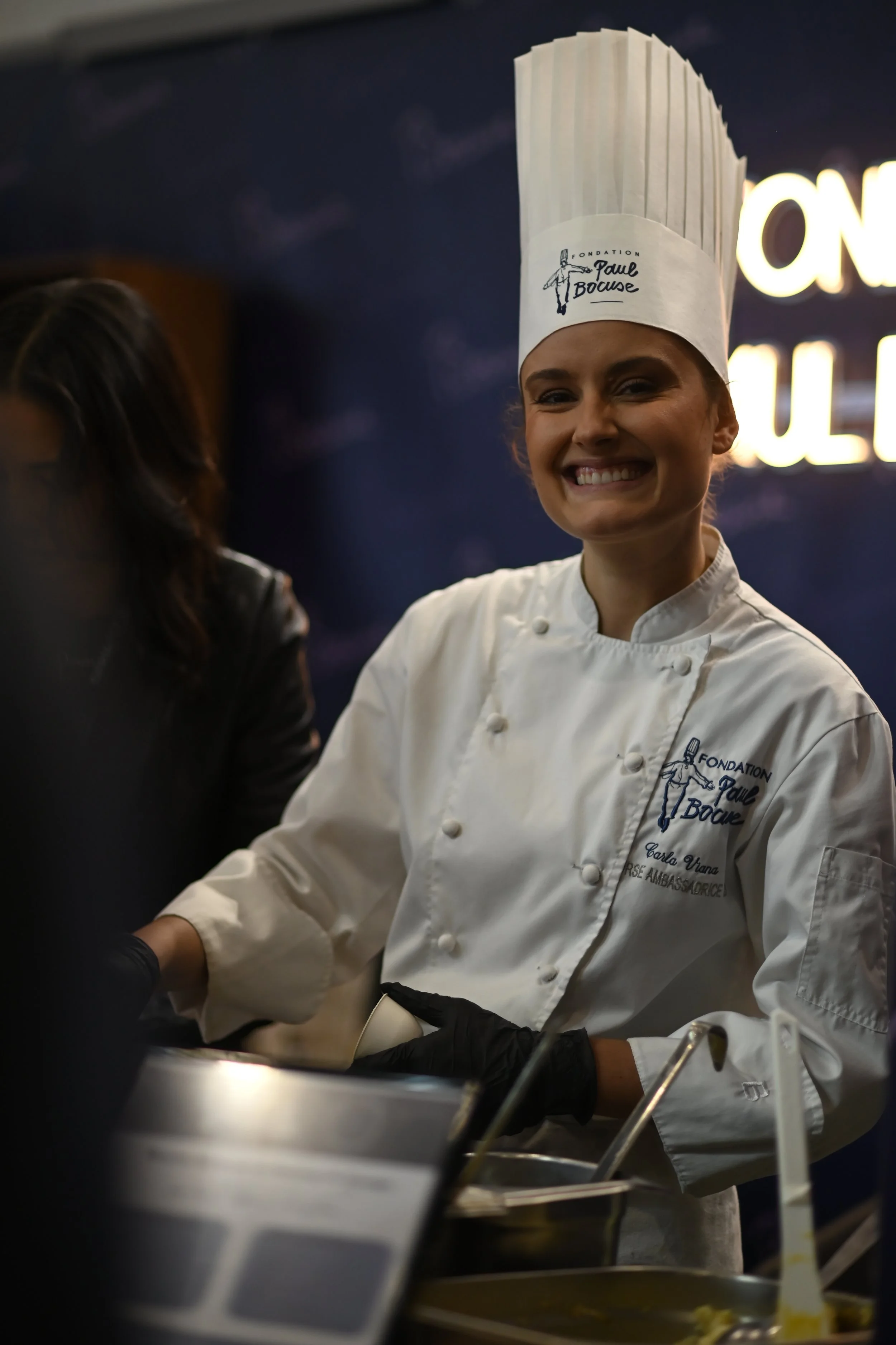 Carla Viana souriante portant une toque blanche avec le logo 'Fondation Paul Bocuse' et un uniforme blanc, en train de préparer un plat dans une cuisine professionnelle.