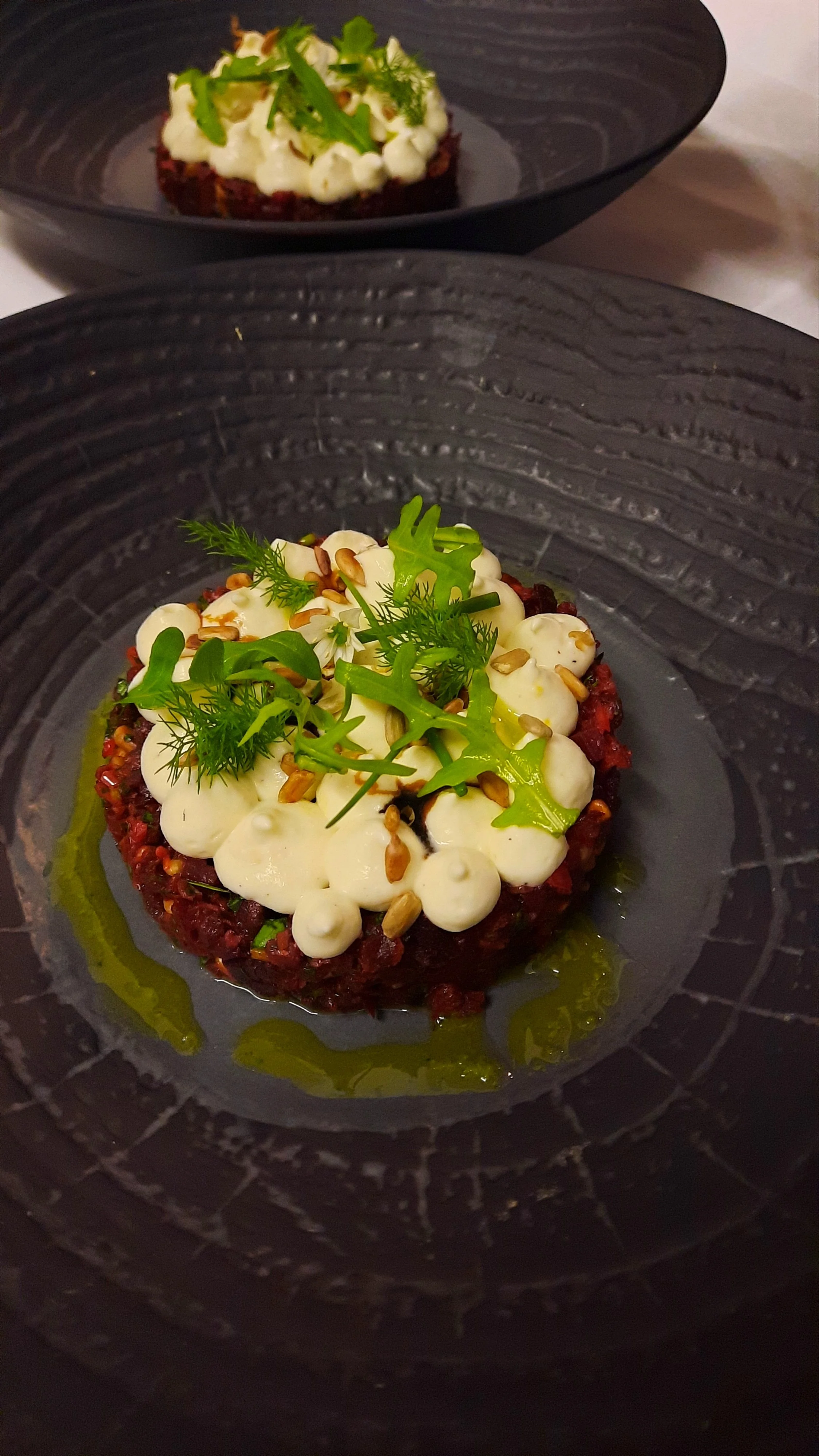 Une assiette de tartare de bœuf garnie de crème, de graines et de légumes verts, présentée sur une assiette noire.