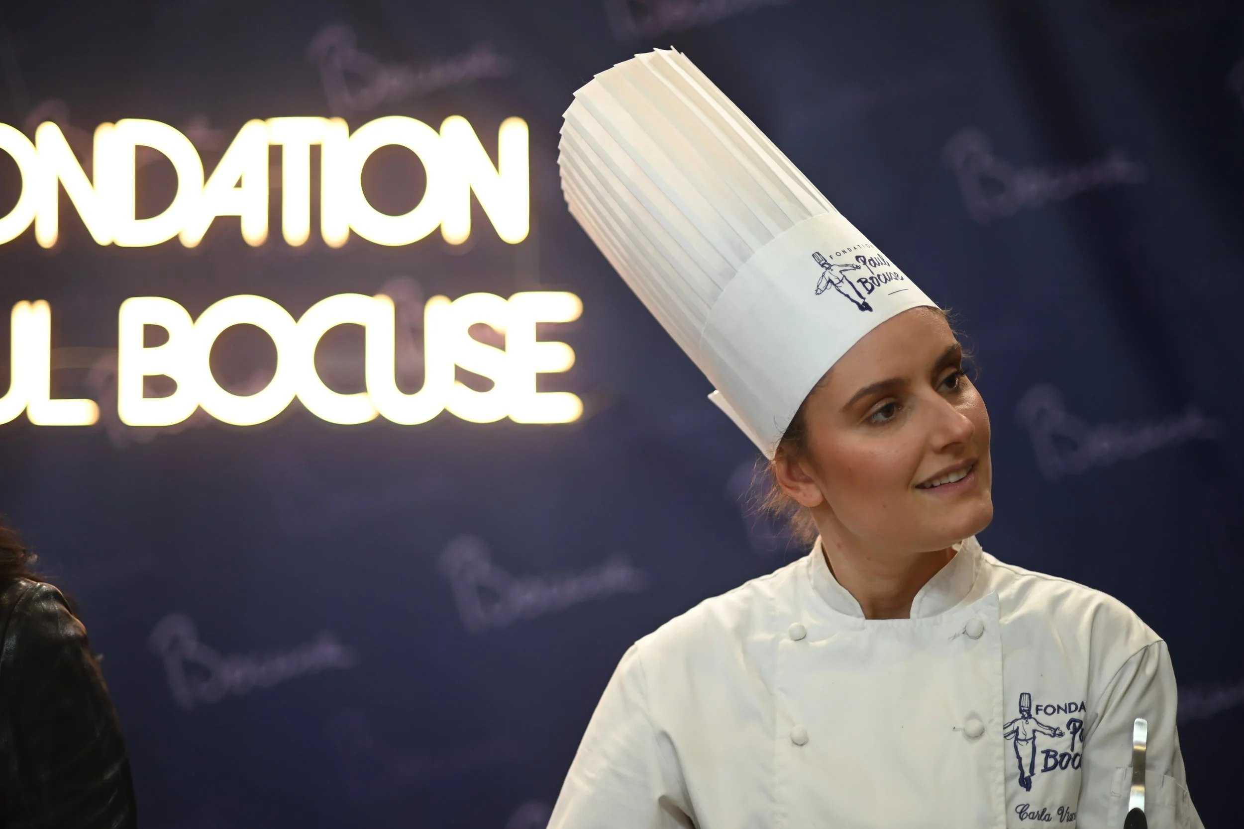 Une femme en uniforme de chef cuisinier porte un chapeau blanc avec un dessin et des inscriptions. Elle sourit, en arrière-plan un panneau lumineux indique 'Fondation du Bonheur'.
