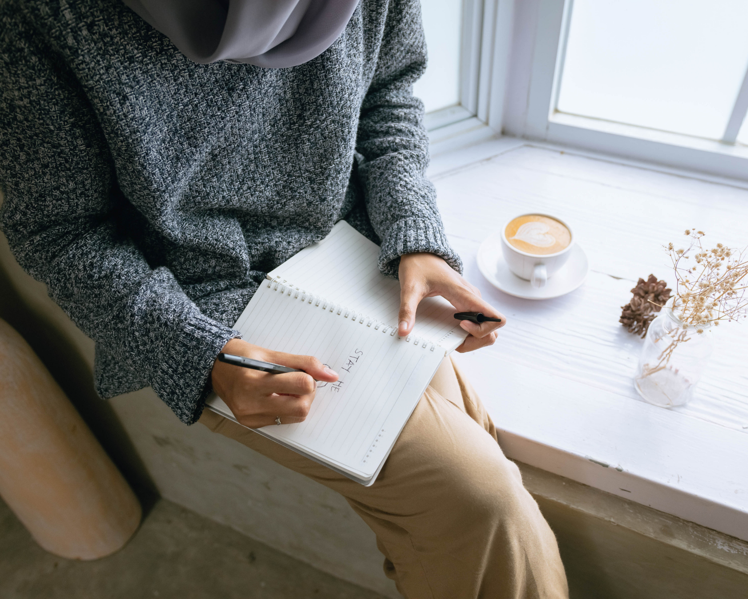 A person sitting on a windowsill writing in a spiral notebook with a pen, with a cup of coffee and a small vase of dried flowers on the sill.