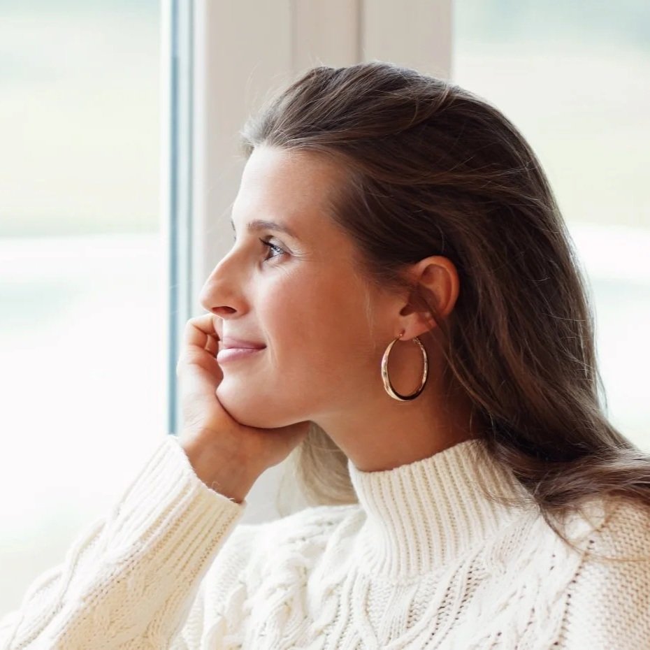 A woman with long brown hair and hoop earrings, looking out a window with a thoughtful expression, wearing a white knitted sweater.