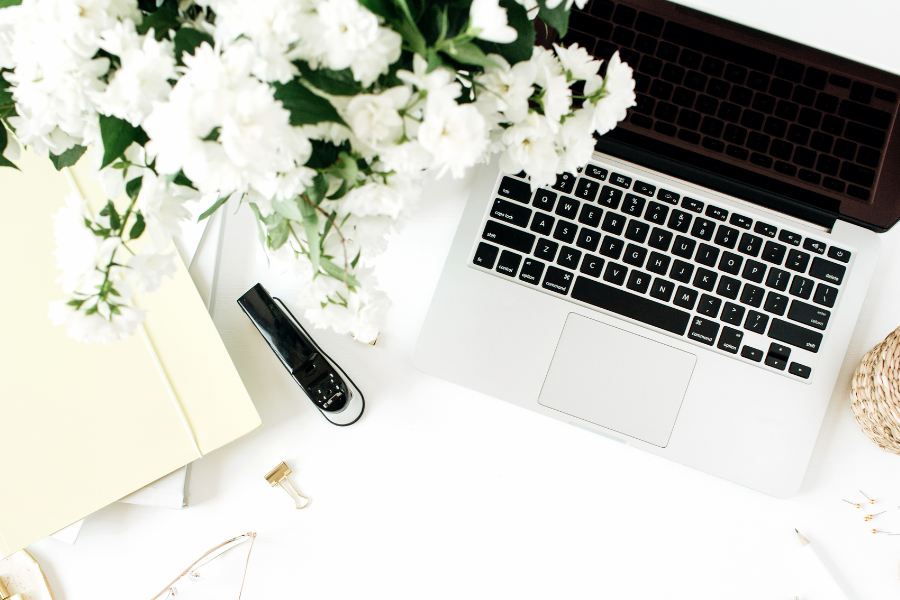 Open laptop with a black screen, white flowers in a vase, stapler, binder clip, and a yellow folder on a white desk.