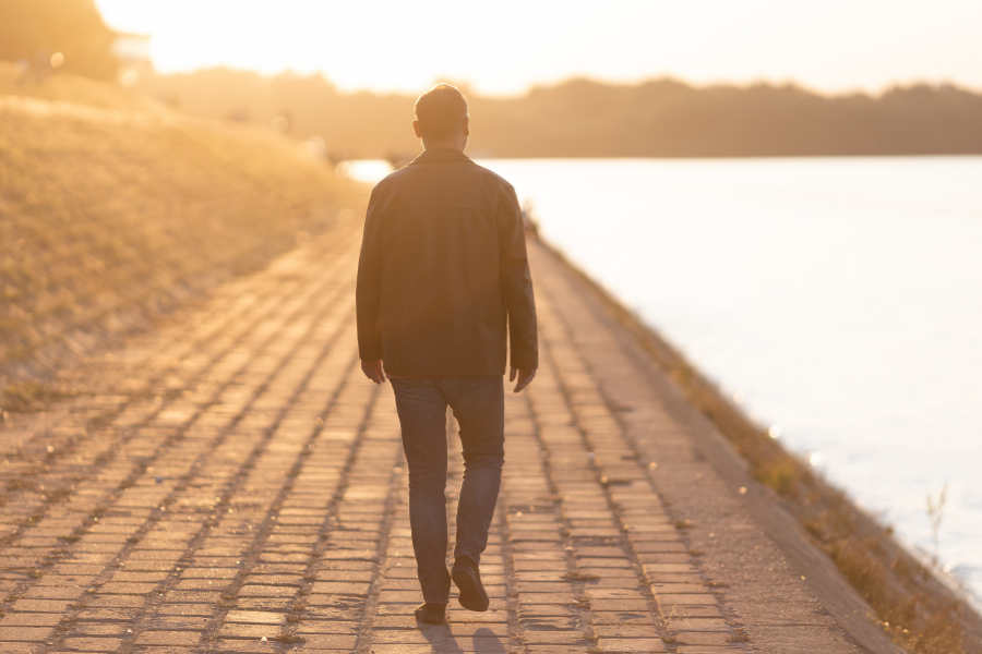 Man walking alone on a paved path by a body of water during sunset.