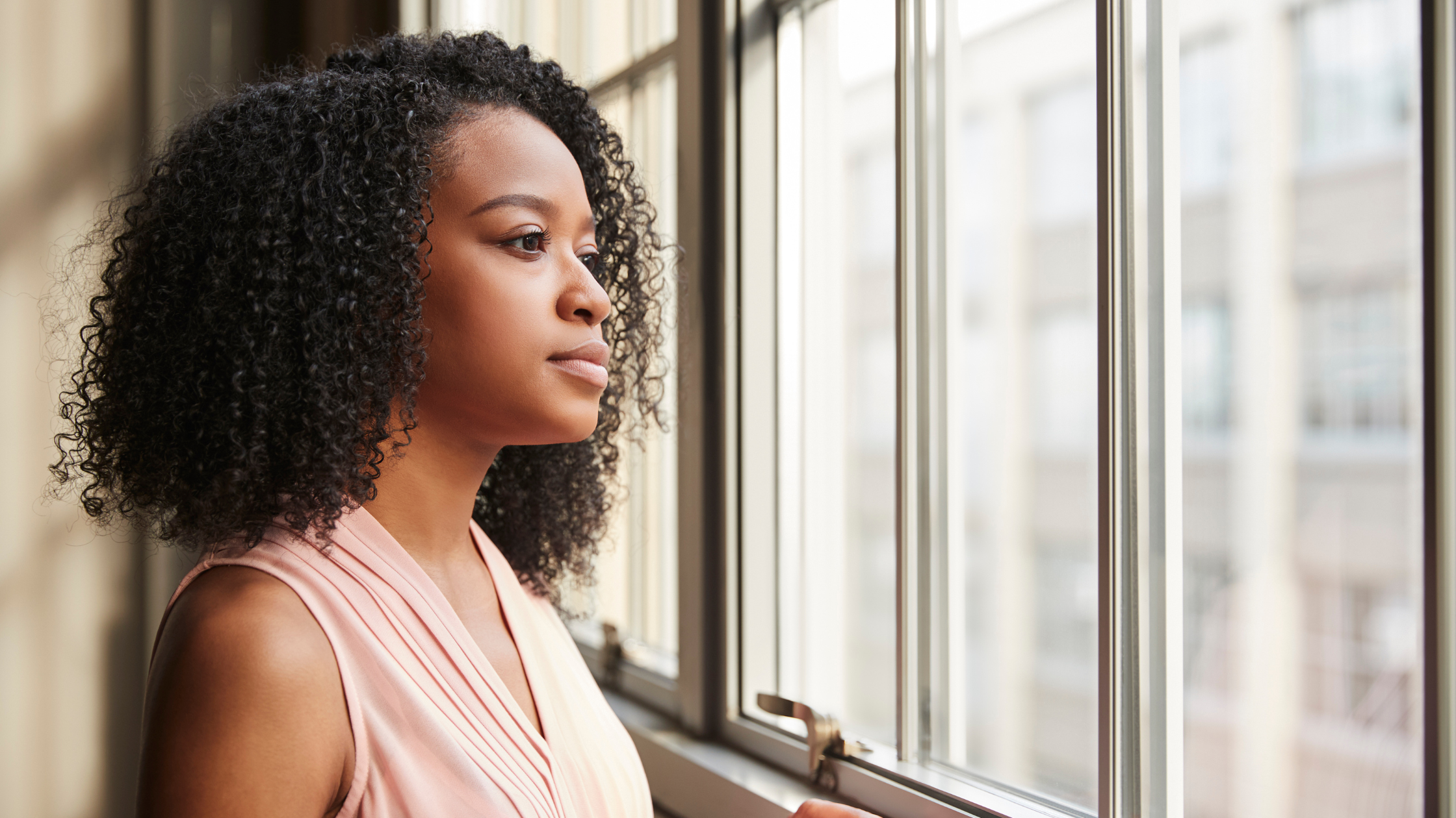 Woman with curly hair looking out a window, wearing a light pink sleeveless top.