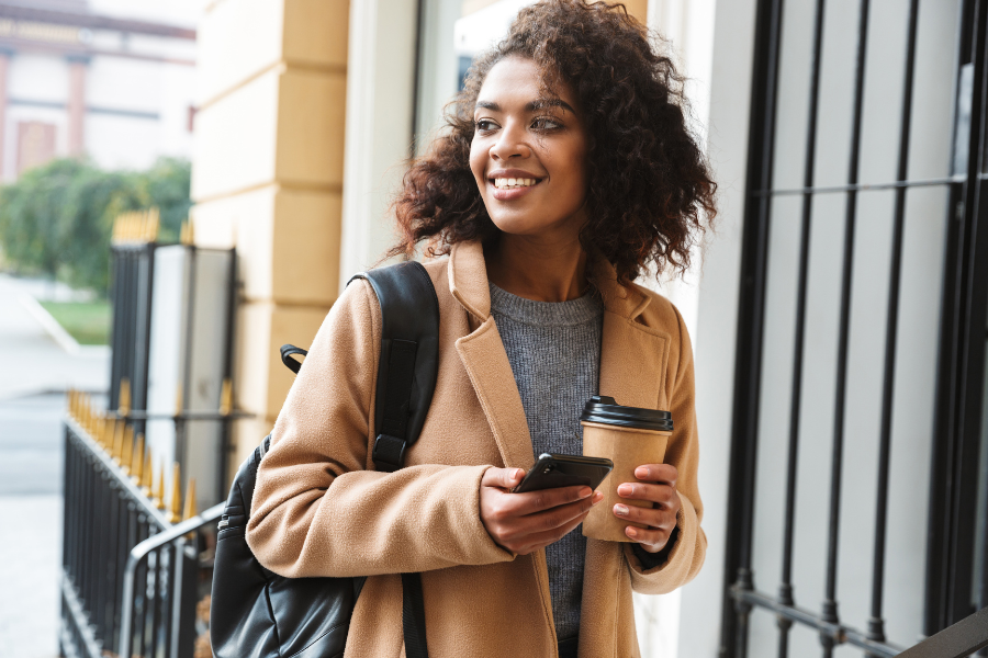 Smiling woman with curly hair holding a smartphone and coffee cup, wearing a tan coat and backpack, standing outdoors.