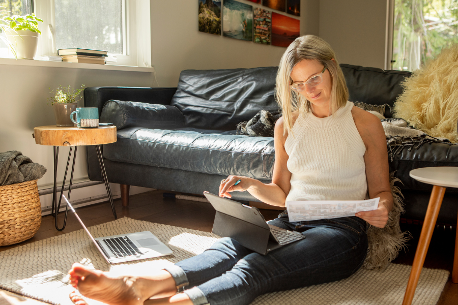 A woman with blonde hair and glasses sitting on the floor in a cozy living room, working on a tablet and holding papers, with sunlight coming through the window.