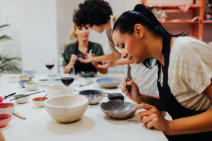 People painting pottery bowls at a table with various supplies and a wine glass.