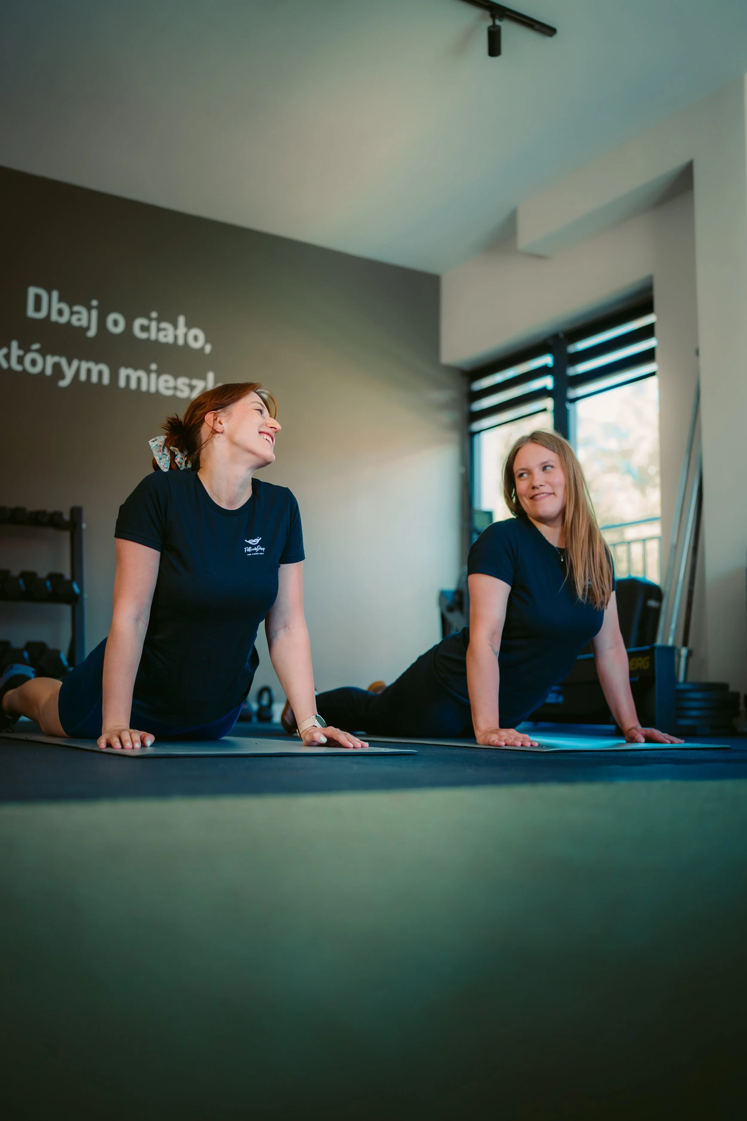 Two women practicing yoga on mats in a gym with large windows, one smiling and the other looking at her, with a dark wall containing text in a foreign language.