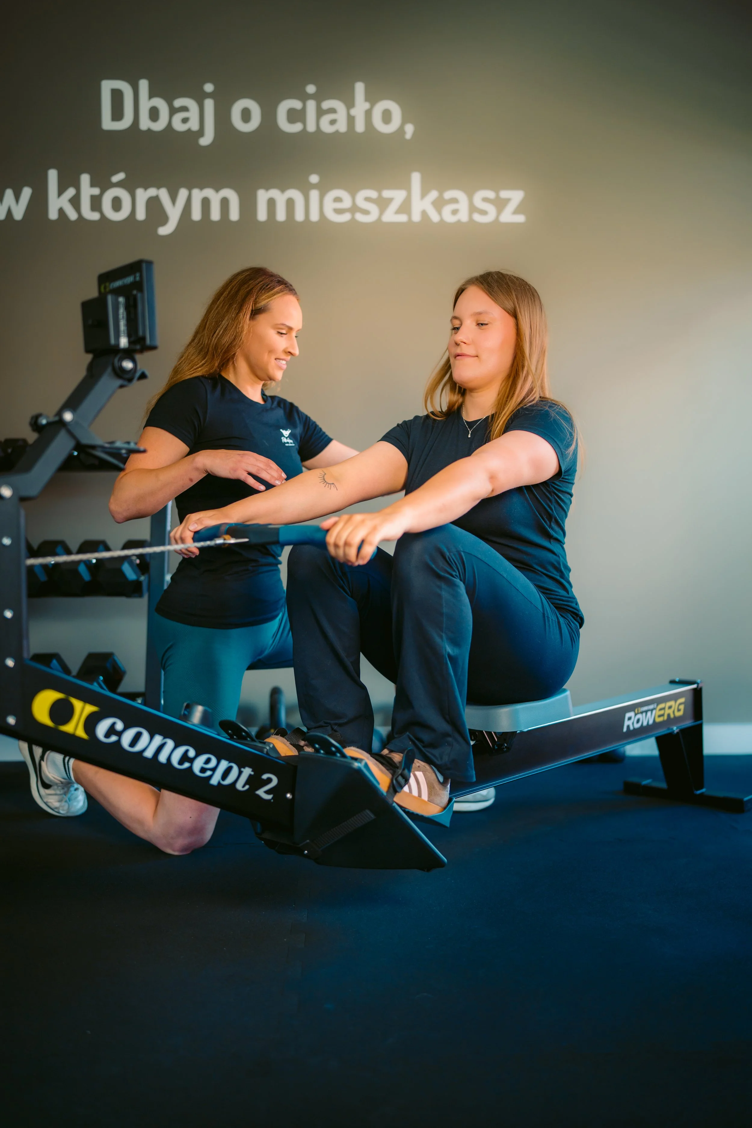 A woman in workout clothing sitting on a rowing machine in a fitness studio, while a trainer suggests her during exercise. The background features a wall with a Polish message about taking care of the body in a healthy environment.