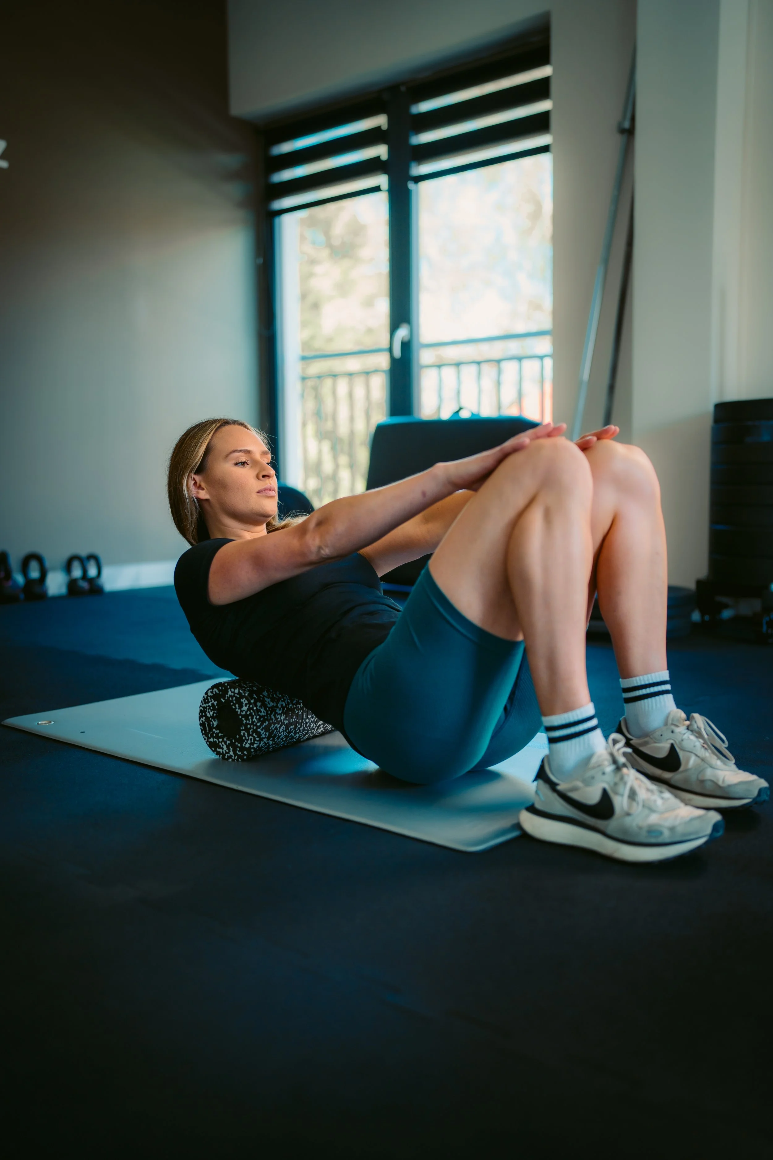 A woman performing sit-up exercise with knees bent, hands clasped behind her head, on a foam roller, wearing workout clothes and sneakers in a gym with large windows and workout equipment in the background.