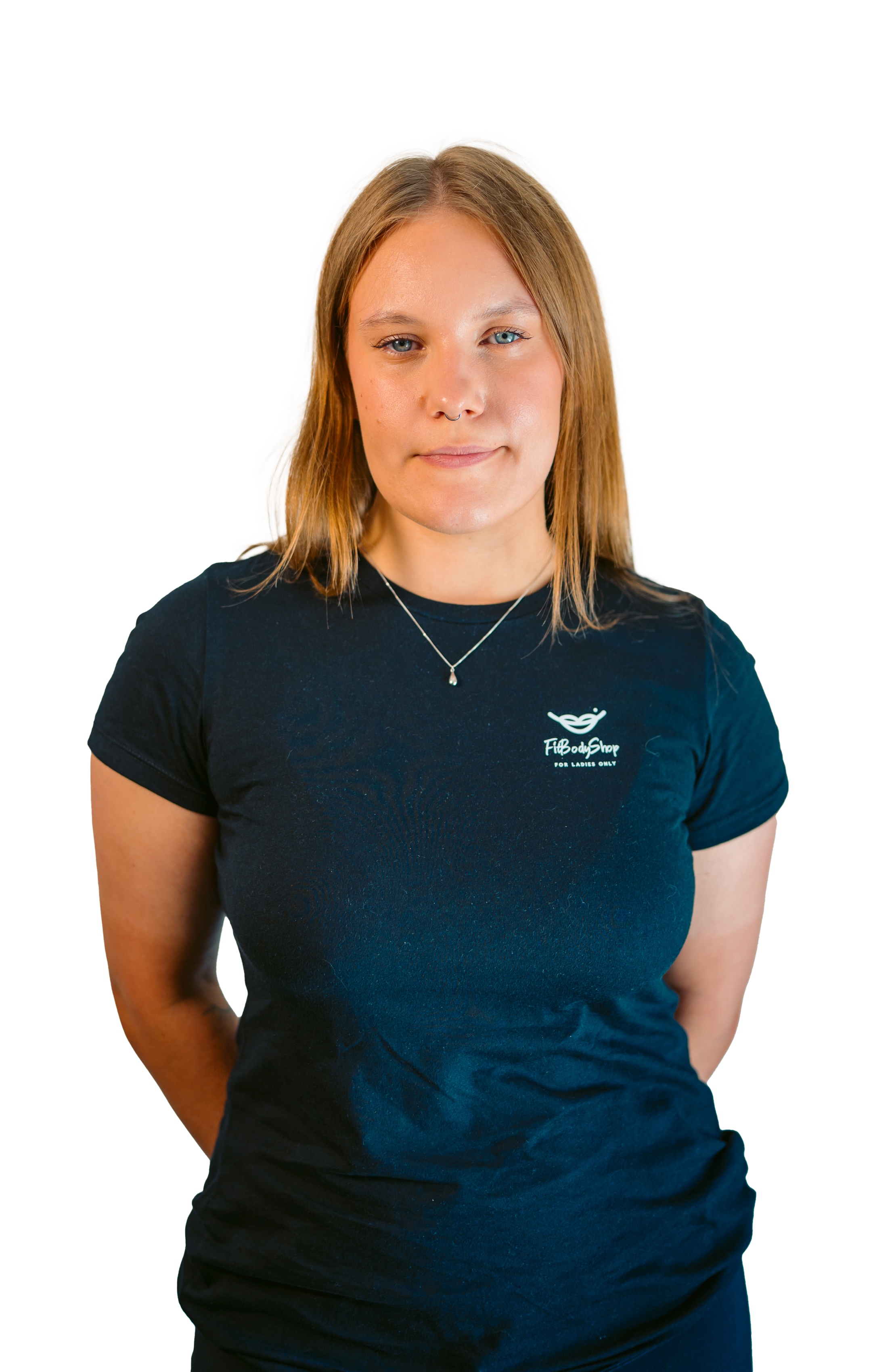 A woman with shoulder-length blonde hair wearing a dark blue t-shirt with a logo on it, standing with her hands behind her back, on a black background.