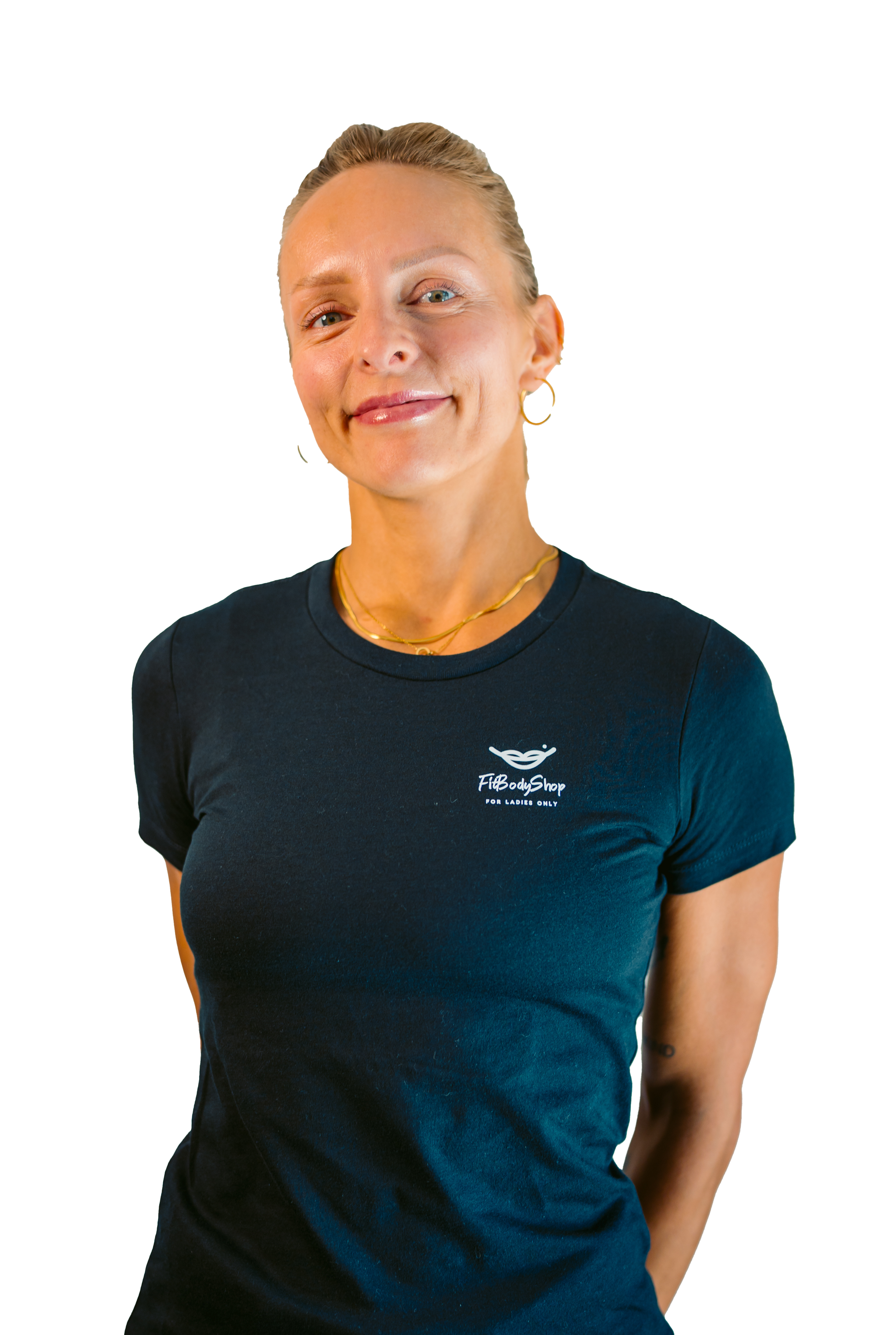A smiling woman with blonde hair tied back, wearing a black t-shirt with the text 'FitBodyShop for Ladies Only' and gold jewelry, standing with her hands behind her back.