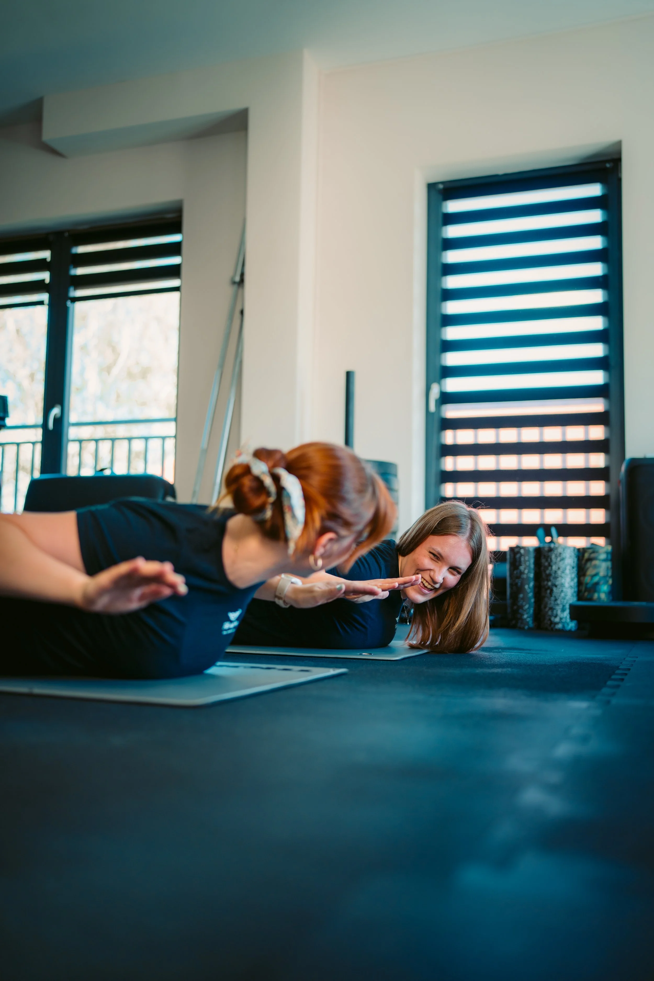 Two women practicing yoga or stretching on mats in a bright room with large windows and black blinds, with a railing outside.