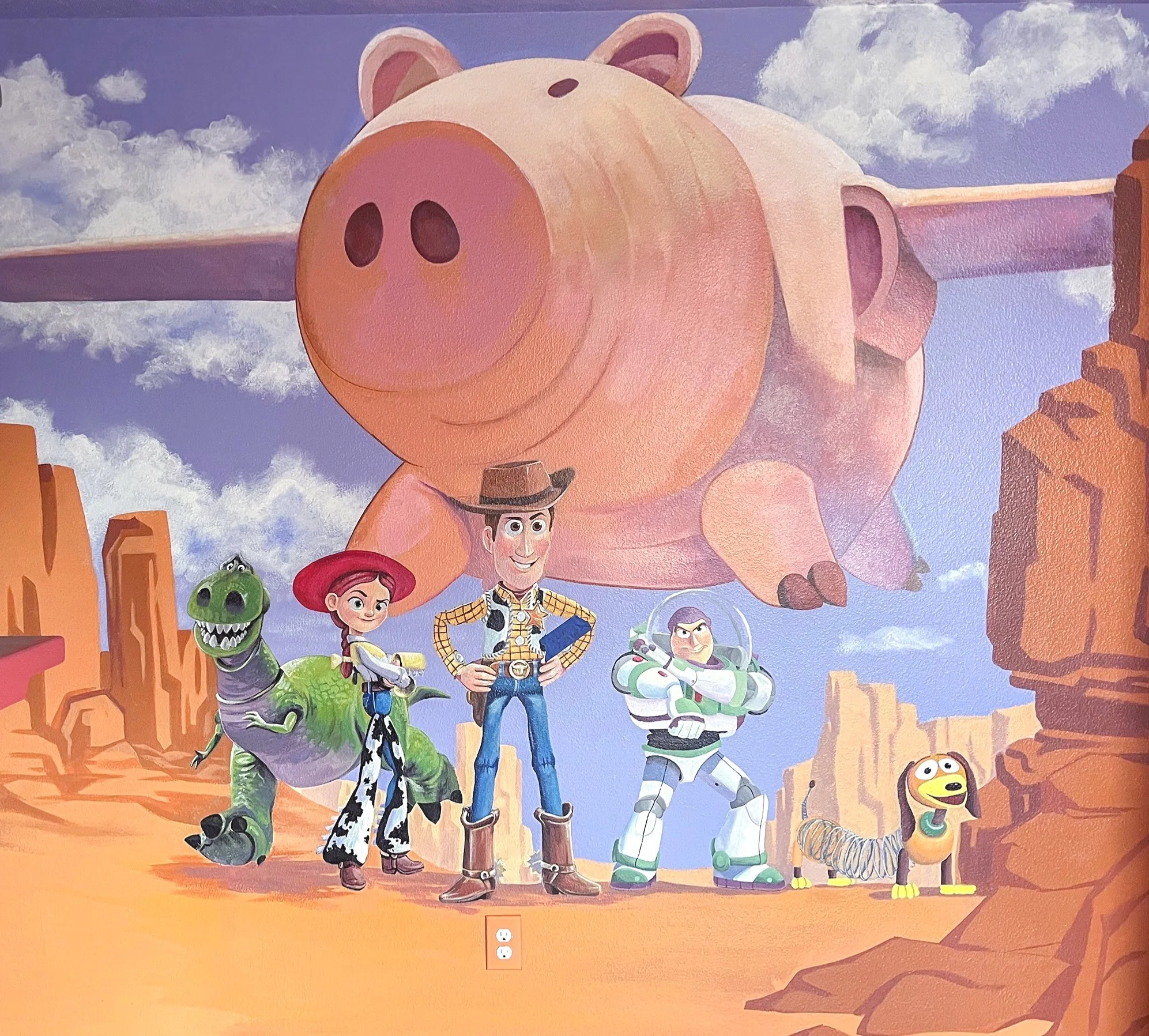 A mural featuring characters from Toy Story, including Woody, Buzz Lightyear, Jessie, Rex, Slinky Dog, and the large pink toy pig from a desert landscape with rock formations and a cloudy sky.
