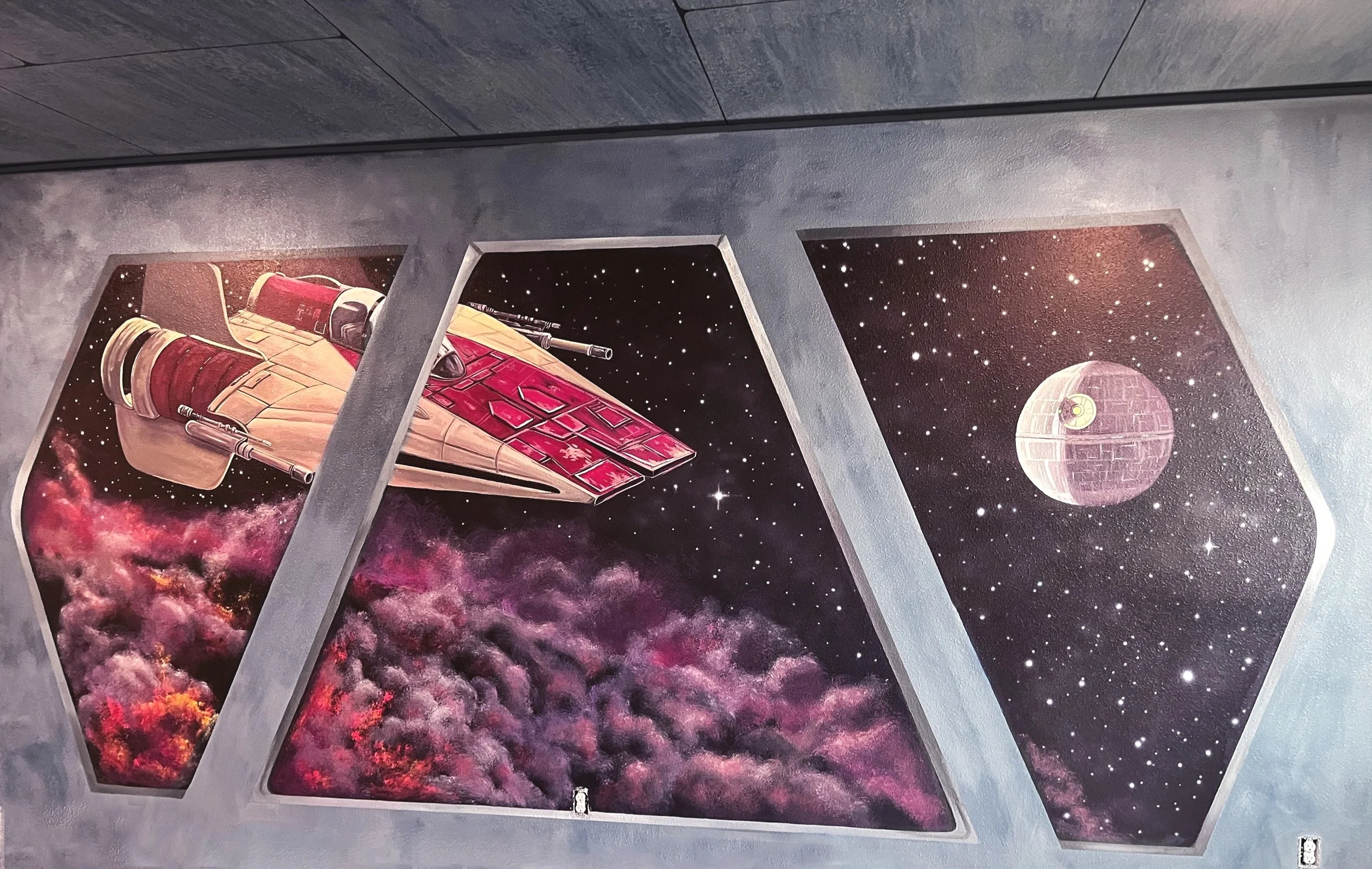 A mural depicting a spaceship near a nebula with pink and purple clouds, and the Death Star from Star Wars in outer space, viewed through three angled windows.