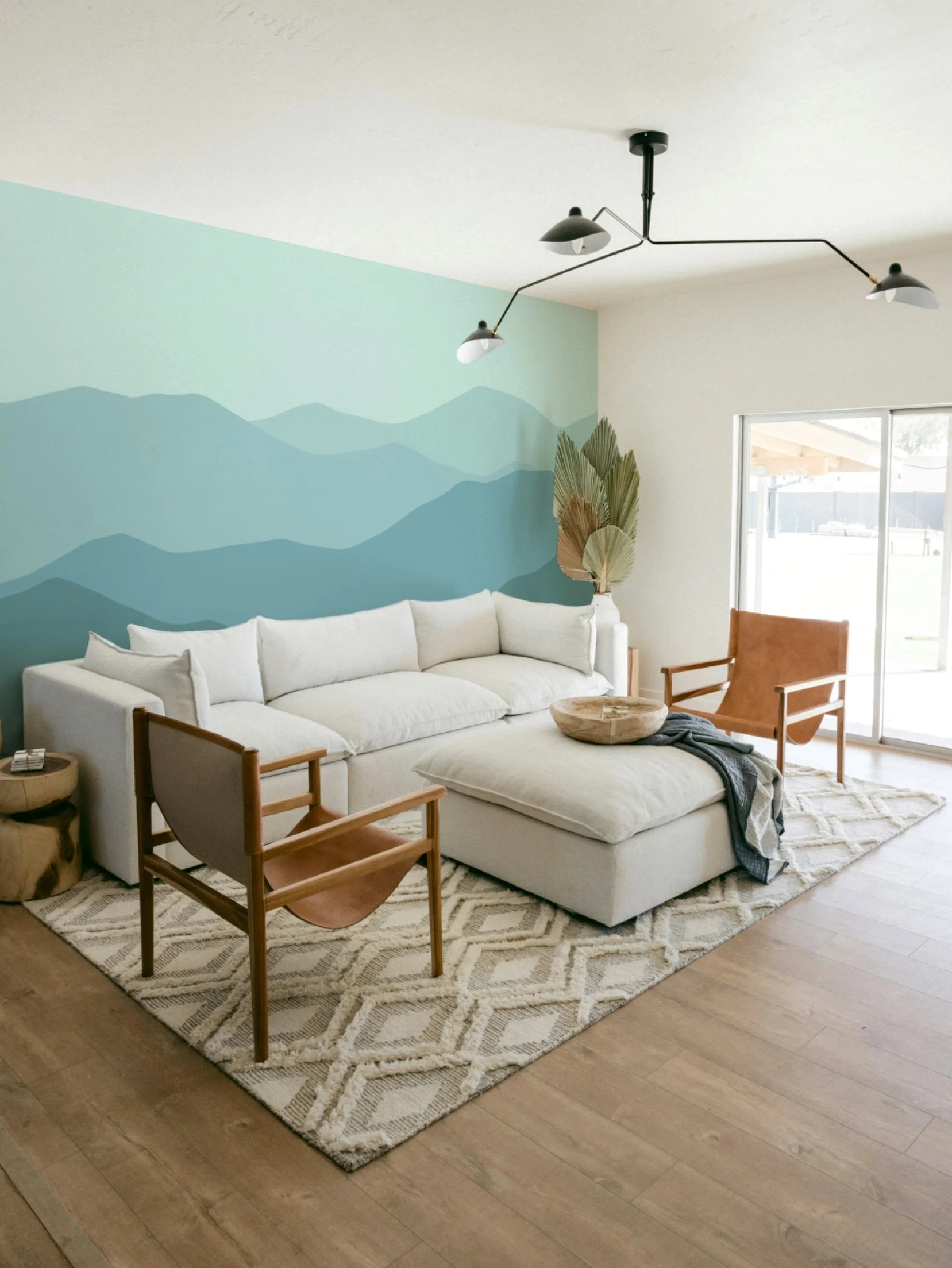 Cozy living room with cream sectional sofa, wooden chairs, a beige rug, and wall art of blue mountains. Large window and modern black chandelier.