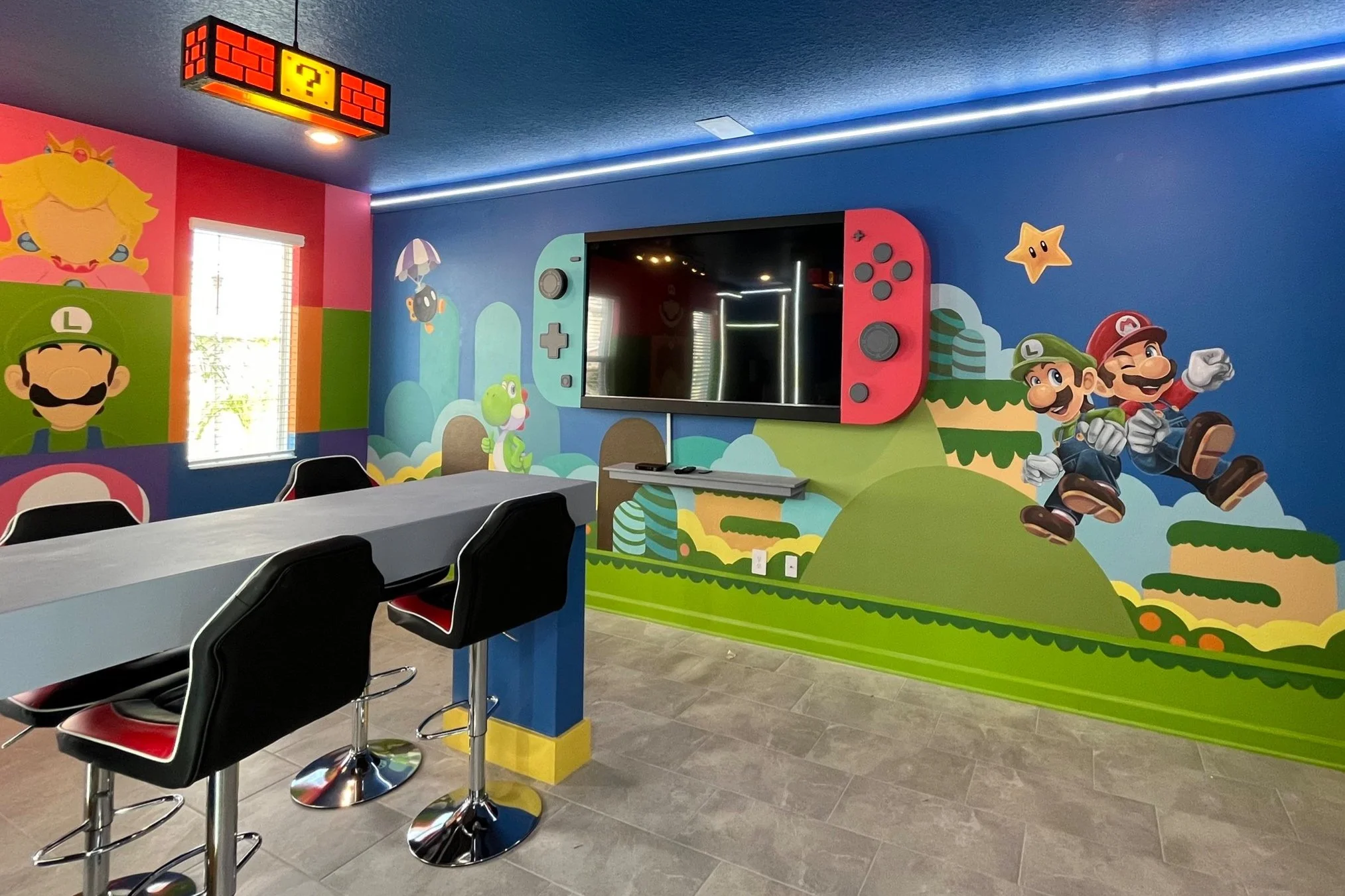Colorful gaming room with Super Mario themed wall mural, featuring Mario and Luigi, Toad, Yoshi, Princess Peach, and various game elements, along with a Nintendo Switch console on the wall, a large TV, and a row of black bar stools.