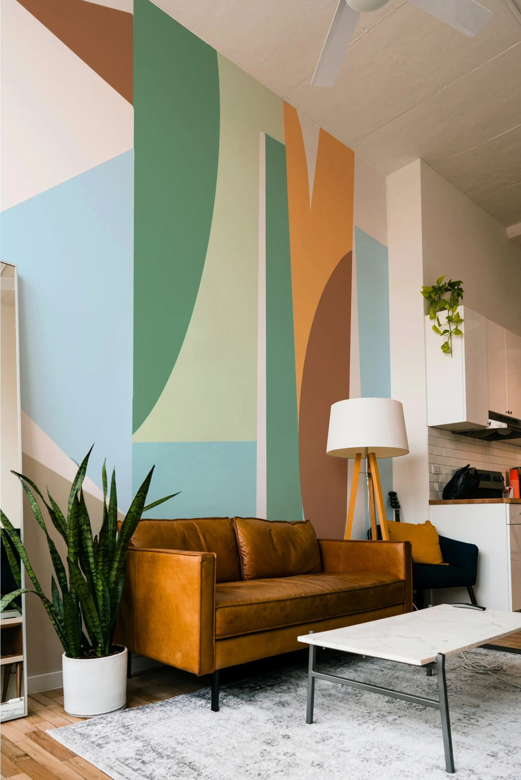 Living room with a brown leather sofa, a tall floor lamp, a large plant in a white pot, a small black wine, a white share, and a colorful abstract mural on the wall.