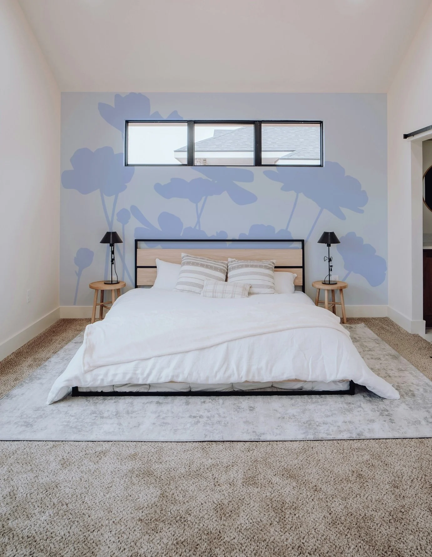 A bedroom with a bed, decorative pillows, two wooden nightstands with black lamps, a light-colored rug, and a wall with a blue floral mural and a horizontal window.
