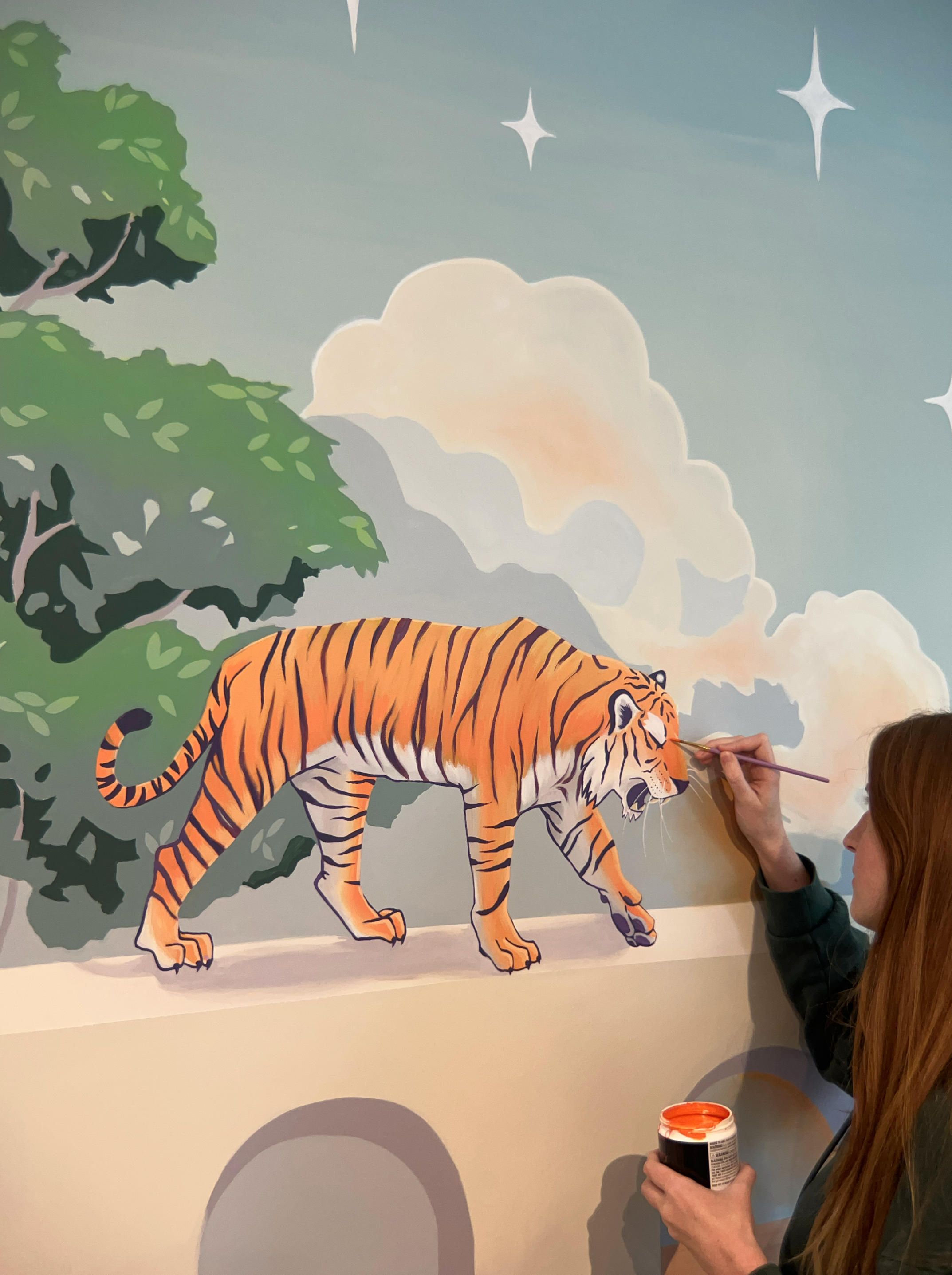 Woman painting a mural of a tiger walking in a jungle scene with trees, clouds, and stars in the background.