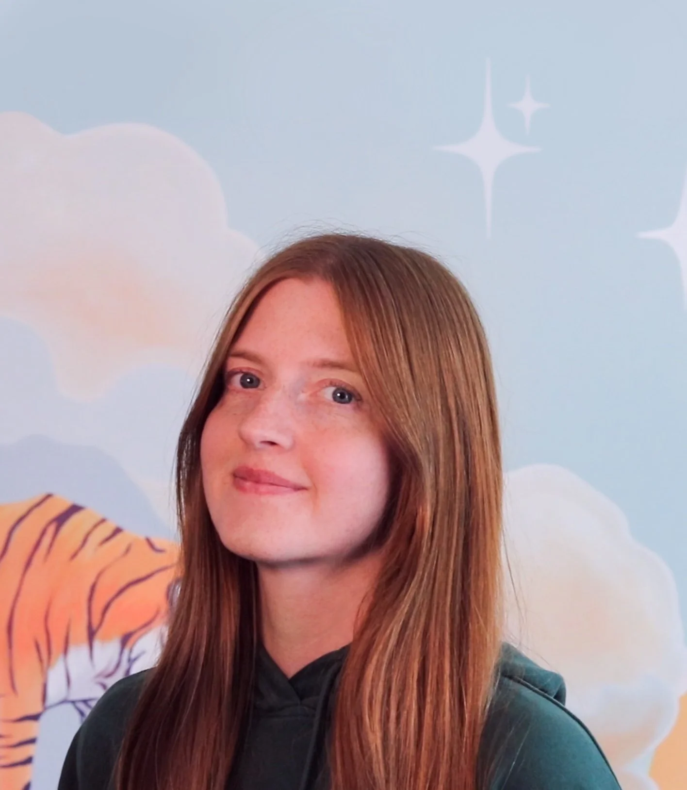 A young woman with long, straight red hair and light skin, smiling softly, wearing a dark hoodie, with a blue sky, clouds, and star accents in the background.