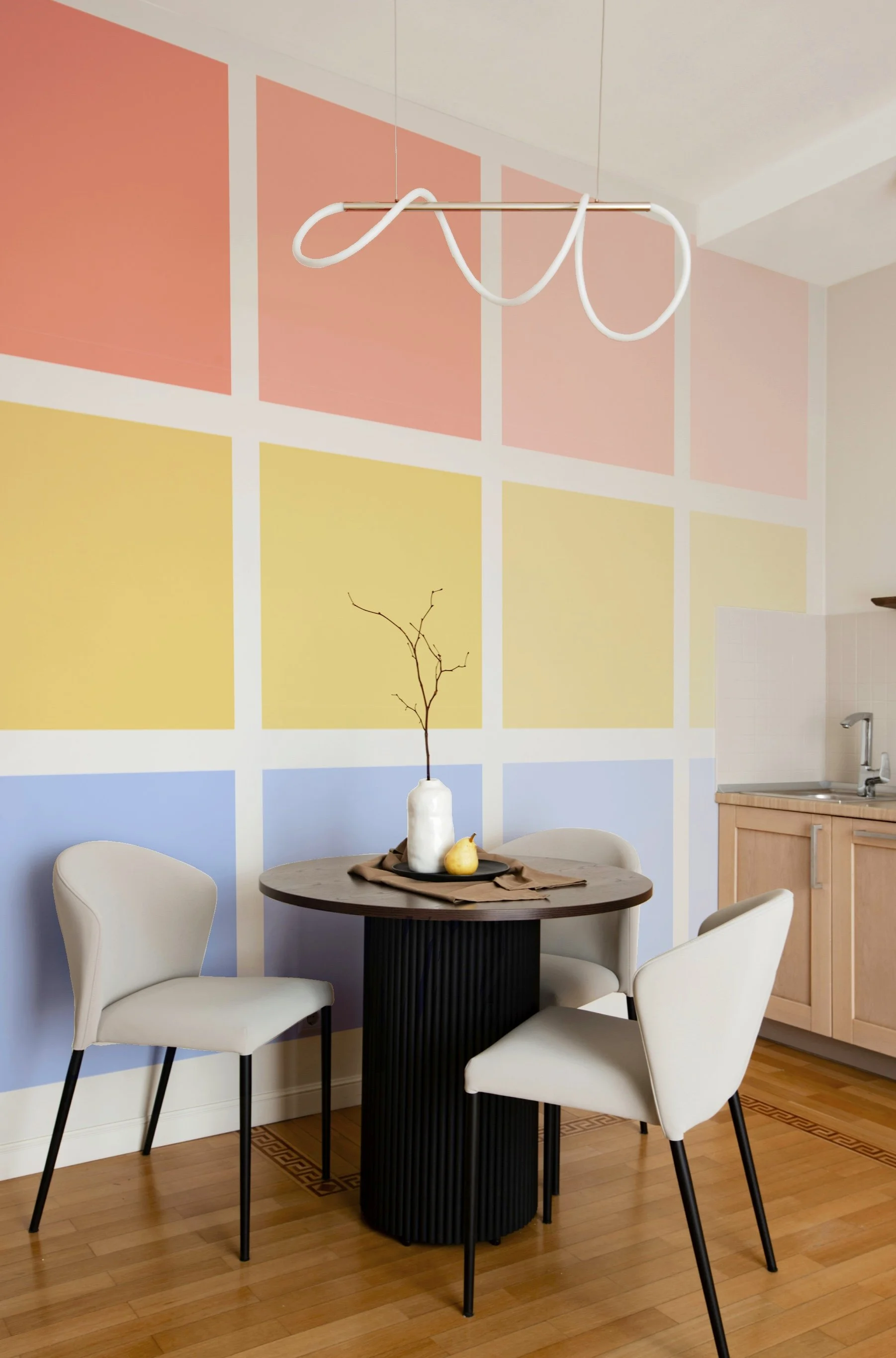 A small dining area with a round black table surrounded by three cream-colored chairs. The table has a white vase with a bare branch and a small pear. The background features a colorful wall with pastel blocks in pink, yellow, and blue, and there is 