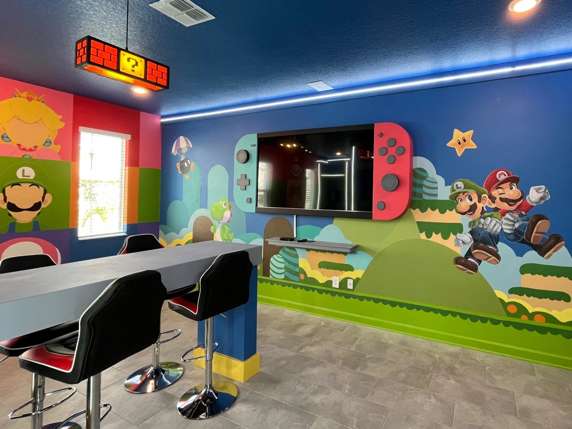 Colorful room decorated with Super Mario themed wall murals, featuring characters like Mario, Luigi, Princess Peach, Yoshi, and Toad, with a modern TV mounted on the wall and a table with chairs.