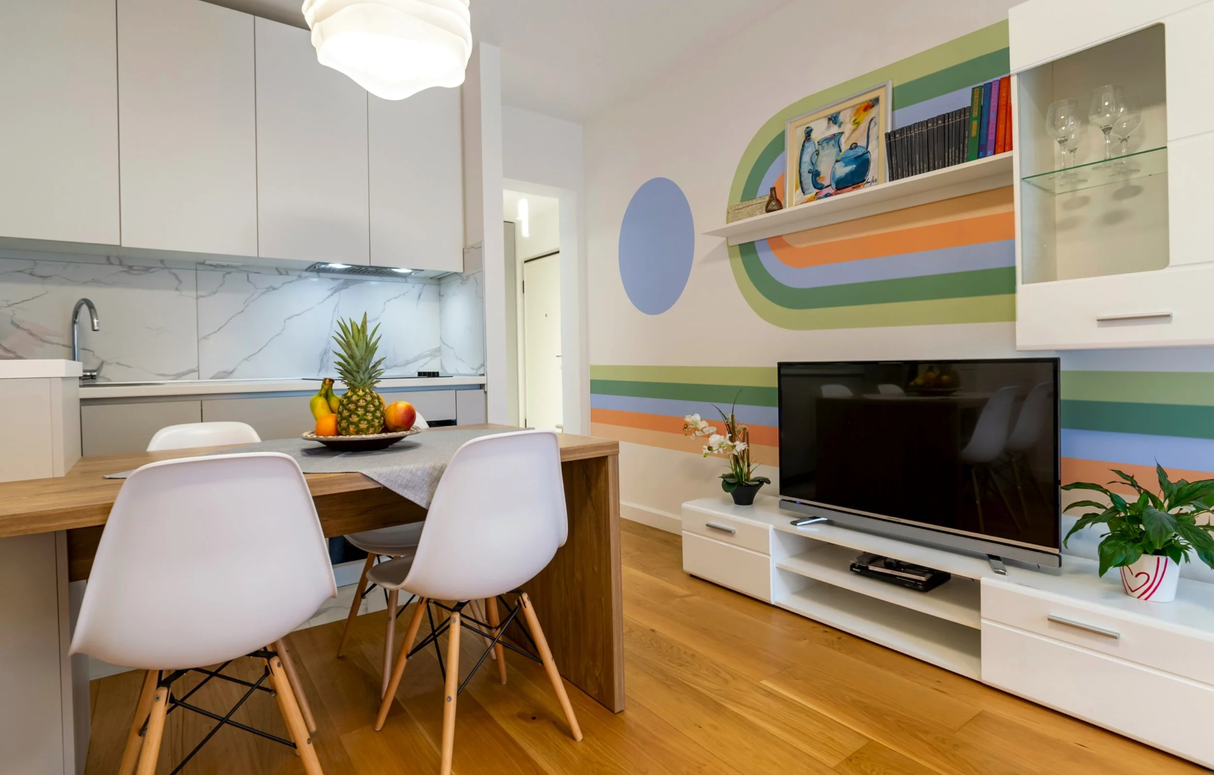 Modern living room and kitchen with colorful wall art, white furniture, a flat-screen TV, a wooden dining table with white chairs, and a bowl of tropical fruits including a pineapple, bananas, and apples.