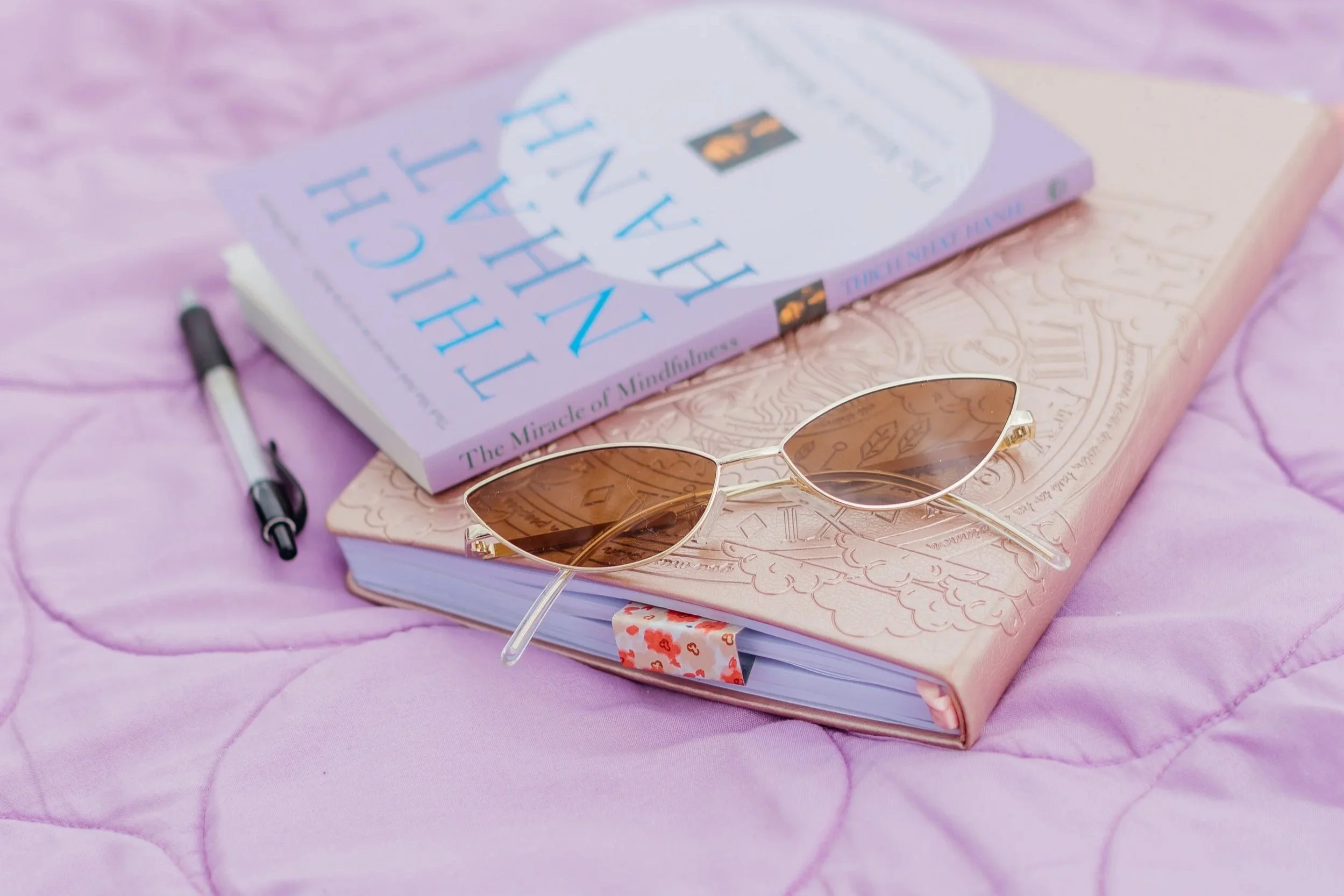 A purple book titled "The Heart Healer" by Frédérique Molay, a pair of sunglasses, and a pen on a pink quilted surface.
