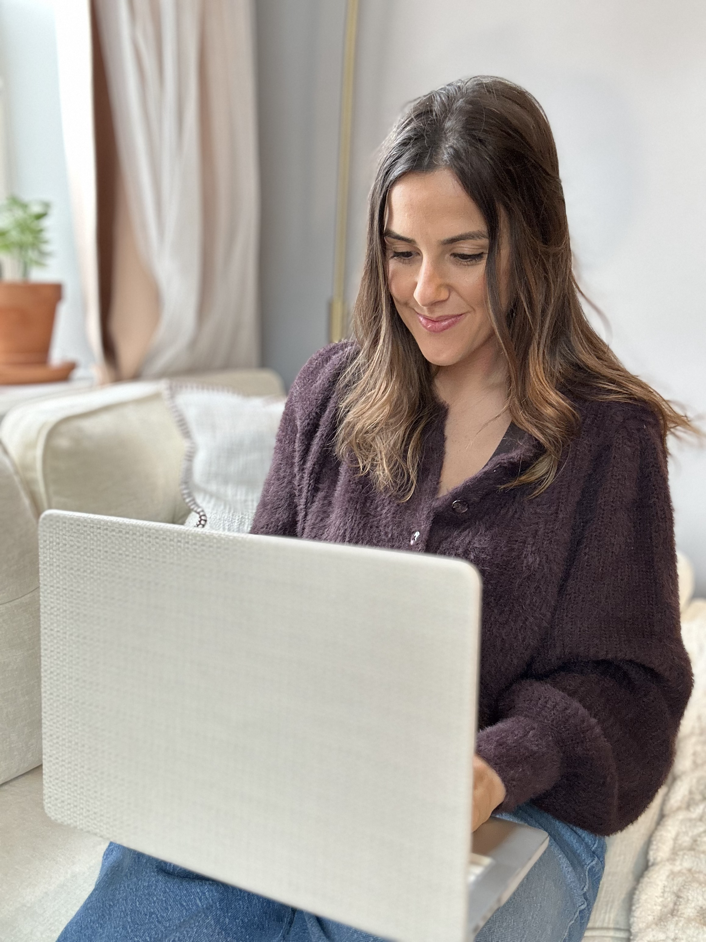 Amanda Pizzo, intuitive reader sitting on a couch using a laptop, with a plant and curtains visible in the background.