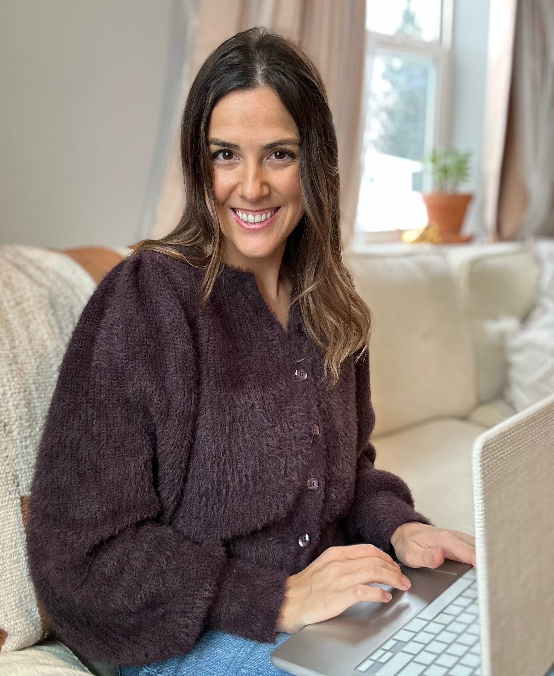Amanda Pizzo, intuitive reading smiling with long brown hair and a warm smile sitting on a beige couch, working on a laptop, in a well-lit room with a window and a potted plant in the background.