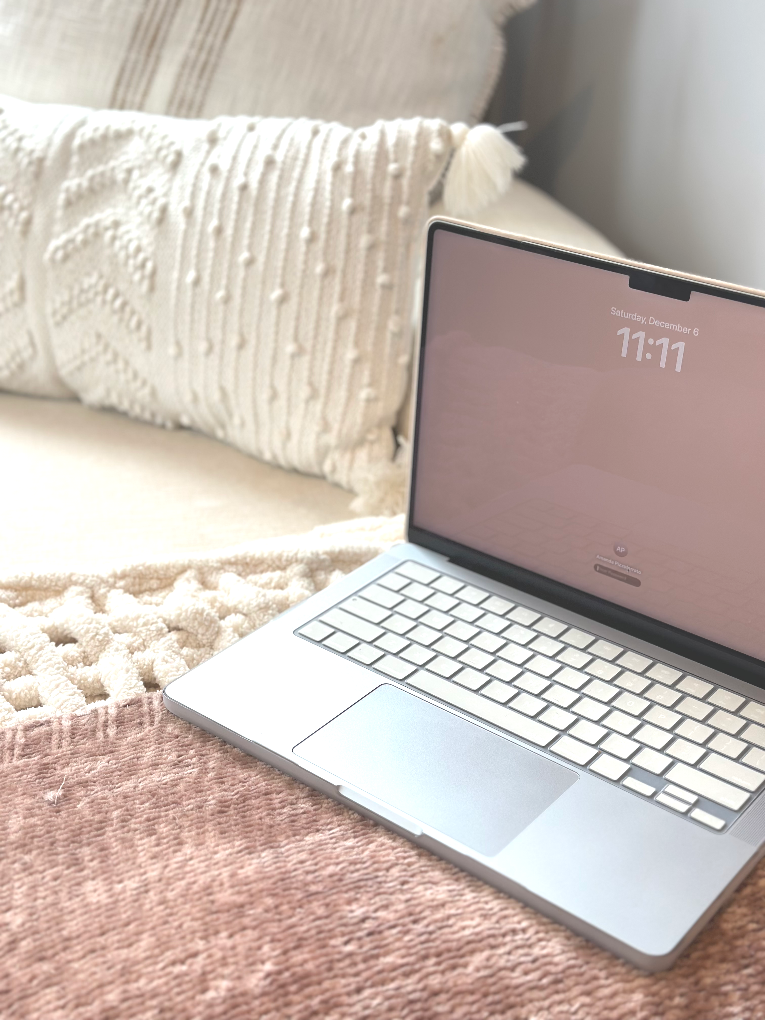Laptop showcasing 11:11 on the screen, on a pink blanket with a white textured pillow and a larger cream-colored pillow in the background.