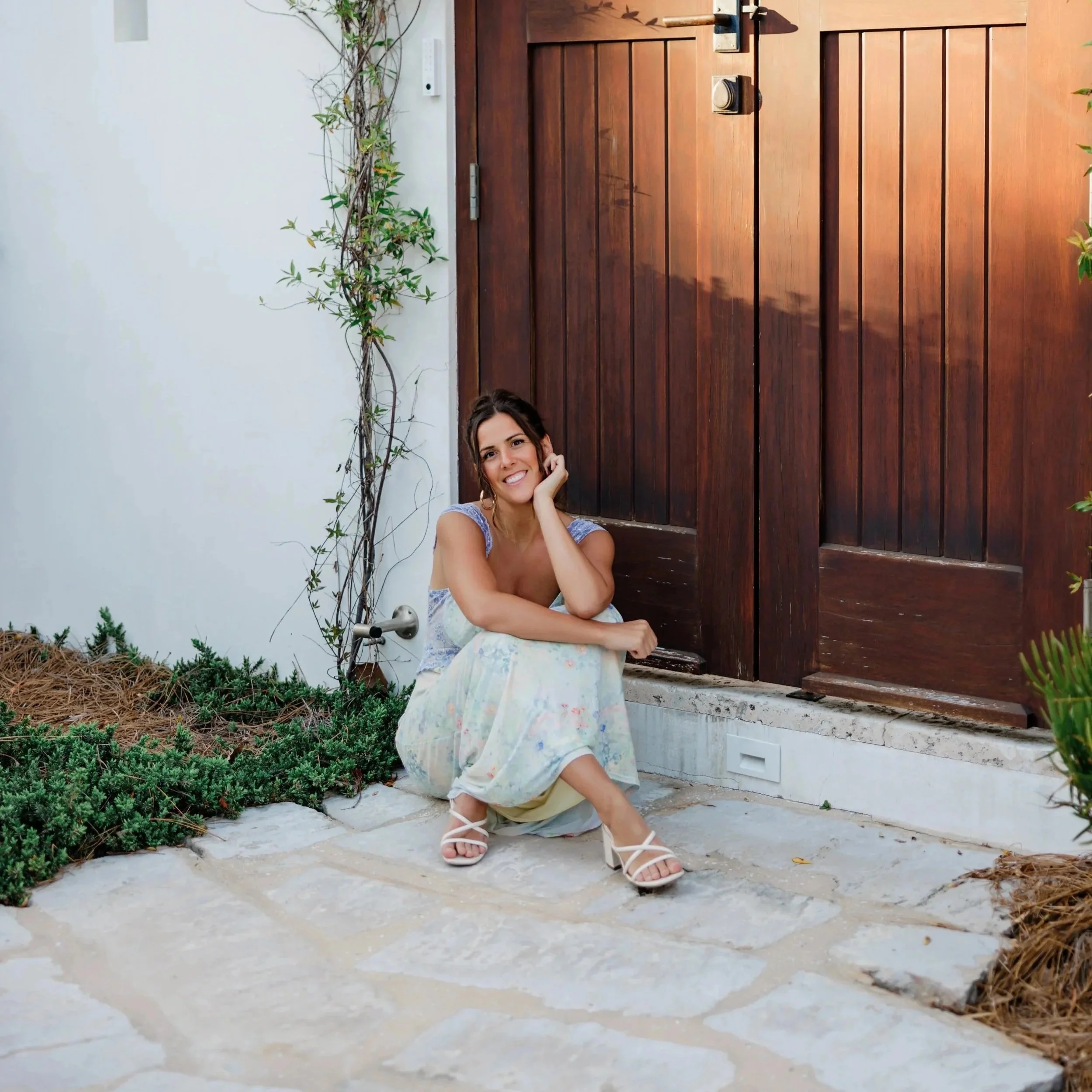 A woman with dark hair sitting on concrete steps in front of a wooden fence and a white wall, smiling and leaning her head on her hand, wearing white sandals and a floral dress.