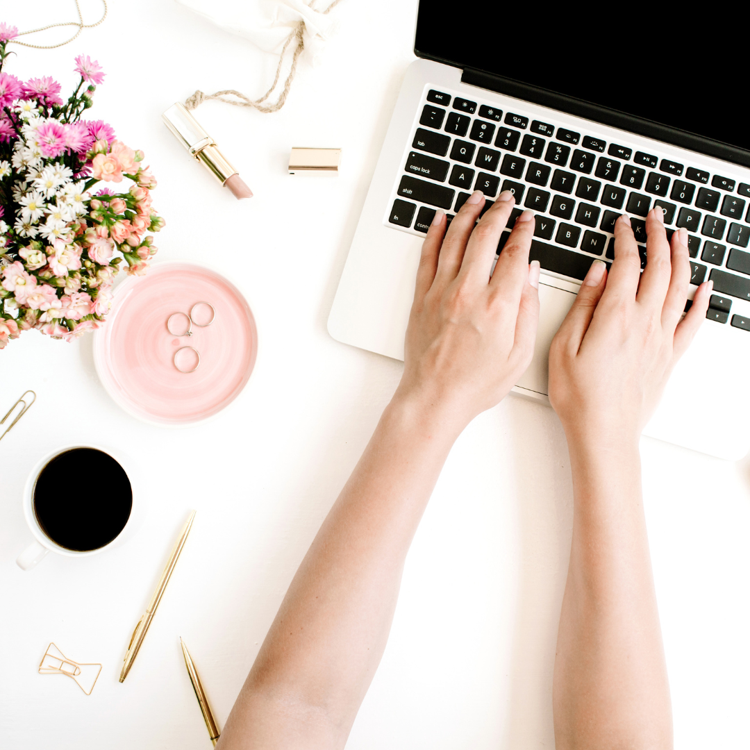A person typing on a laptop with hands placed on the keyboard, surrounded by a cup of coffee, a pink plate with rings, a tube of lipstick, a bouquet of pink and white flowers, a gold paperclip, gold pens, and a small jewelry box on a white desk.