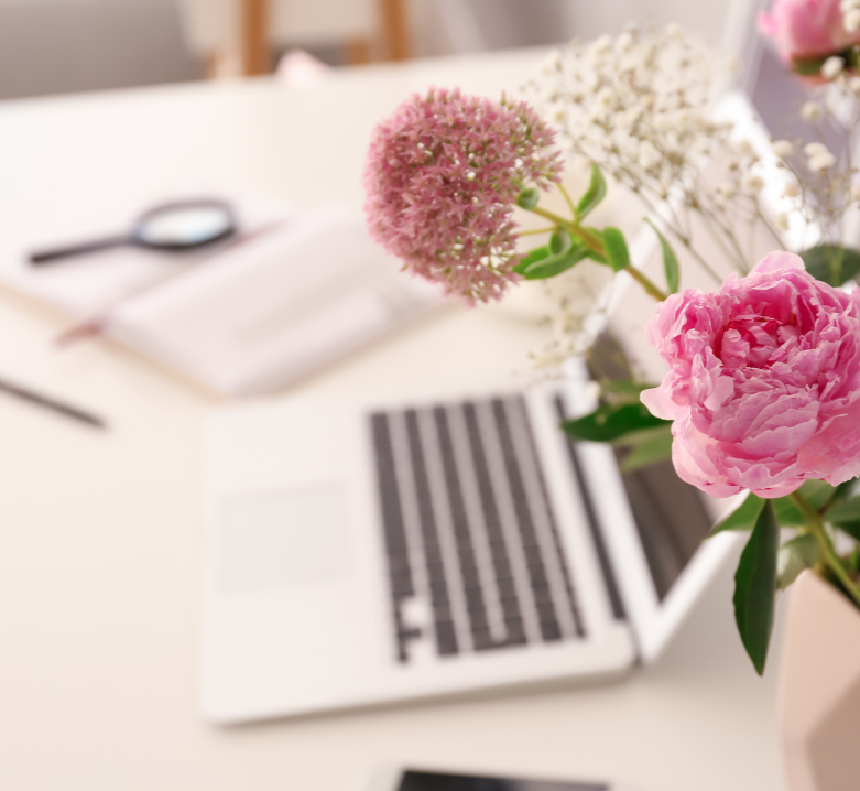 Close-up of a laptop and workspace with pink and white flowers in a vase on a white desk.