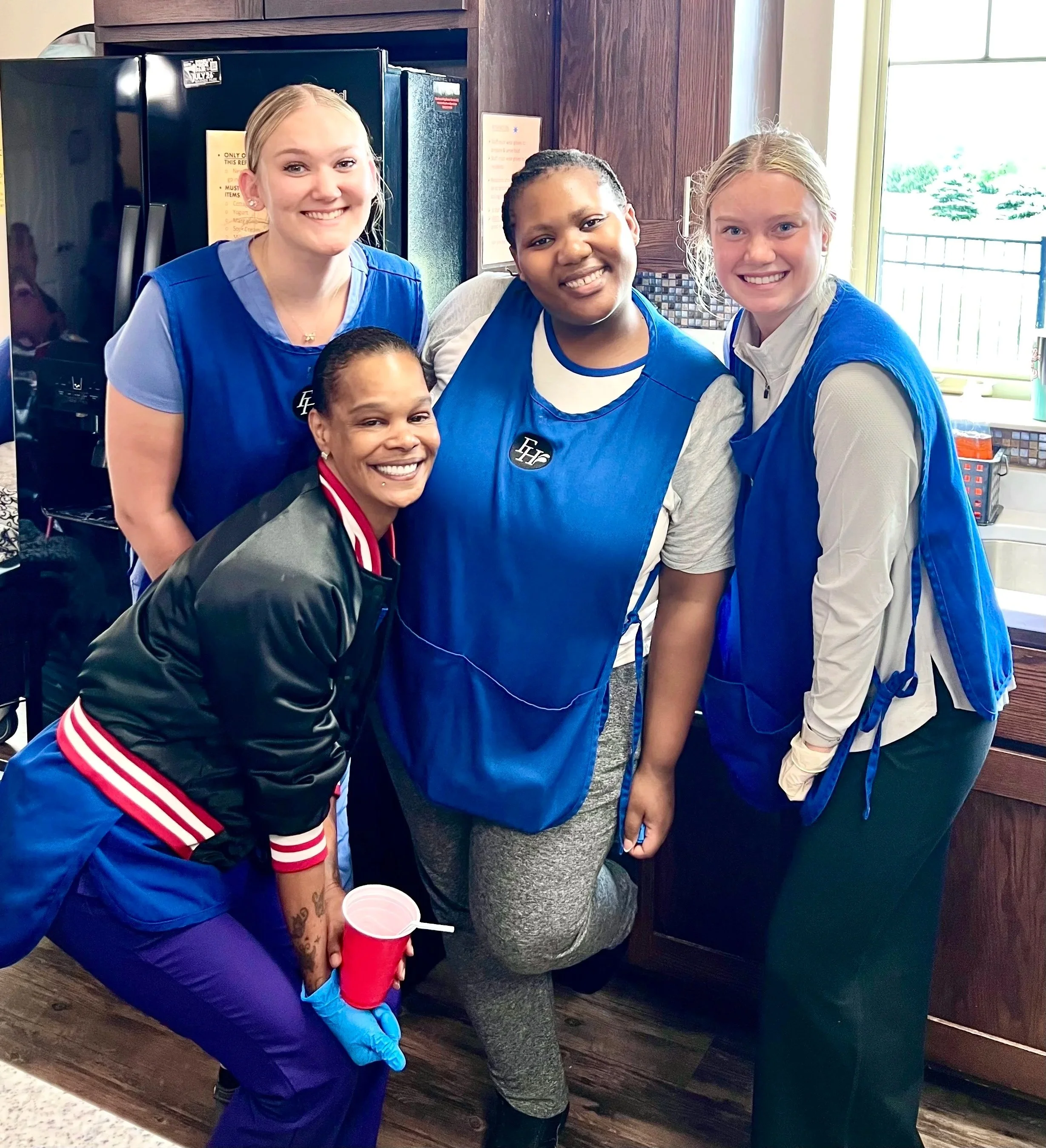 A group of women 4 Fountainhead Homes employees, women, standing together in taking a photo in the kitchen of one of the facilities. They are all smiling in a group photo showing the joy and laughter they hav while working at Fountainhead Homes.