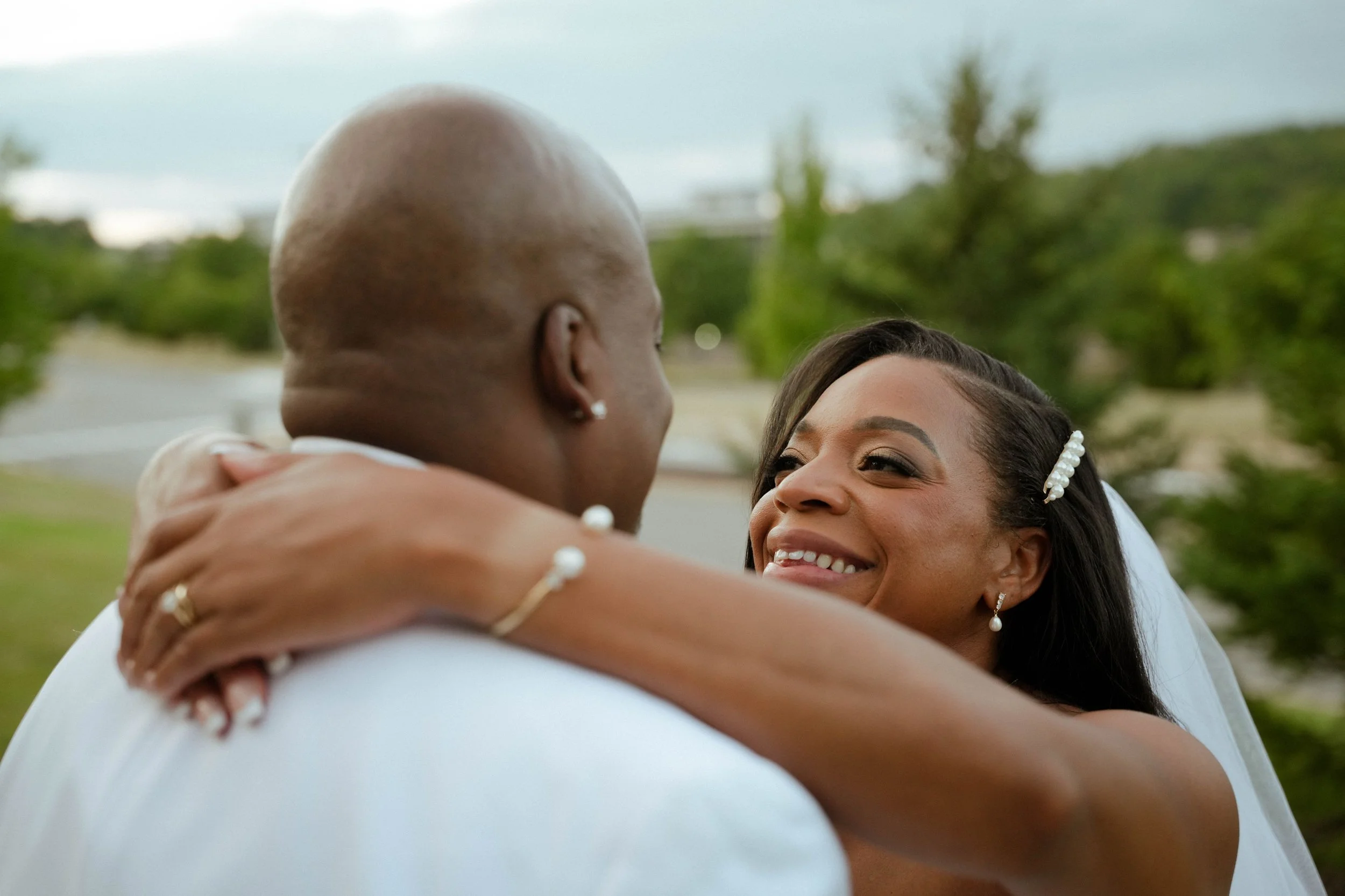 A joyful couple embracing outdoors, with the woman smiling at the man. The woman has dark hair, a pearl hair accessory, and pearl earrings. The man has a bald head and earrings, and is wearing a white shirt. Green trees and cloudy sky are in the back