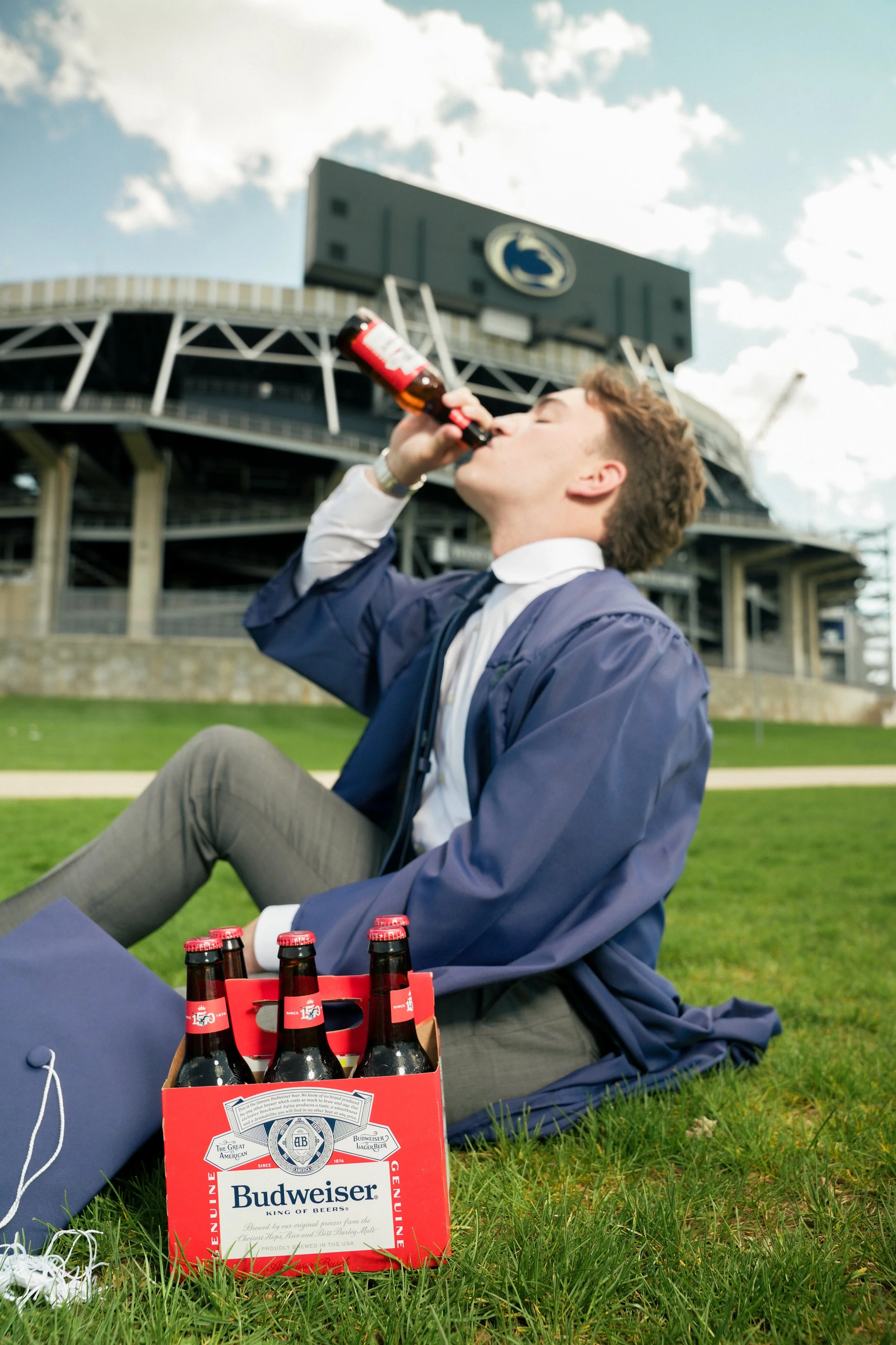 A young man in a blue jacket and white shirt sitting on grass in front of a baseball stadium, drinking from a bottle. A six-pack of Budweiser beer is on the grass nearby, with a red beer carrier in front of him. The stadium has a modern design with a