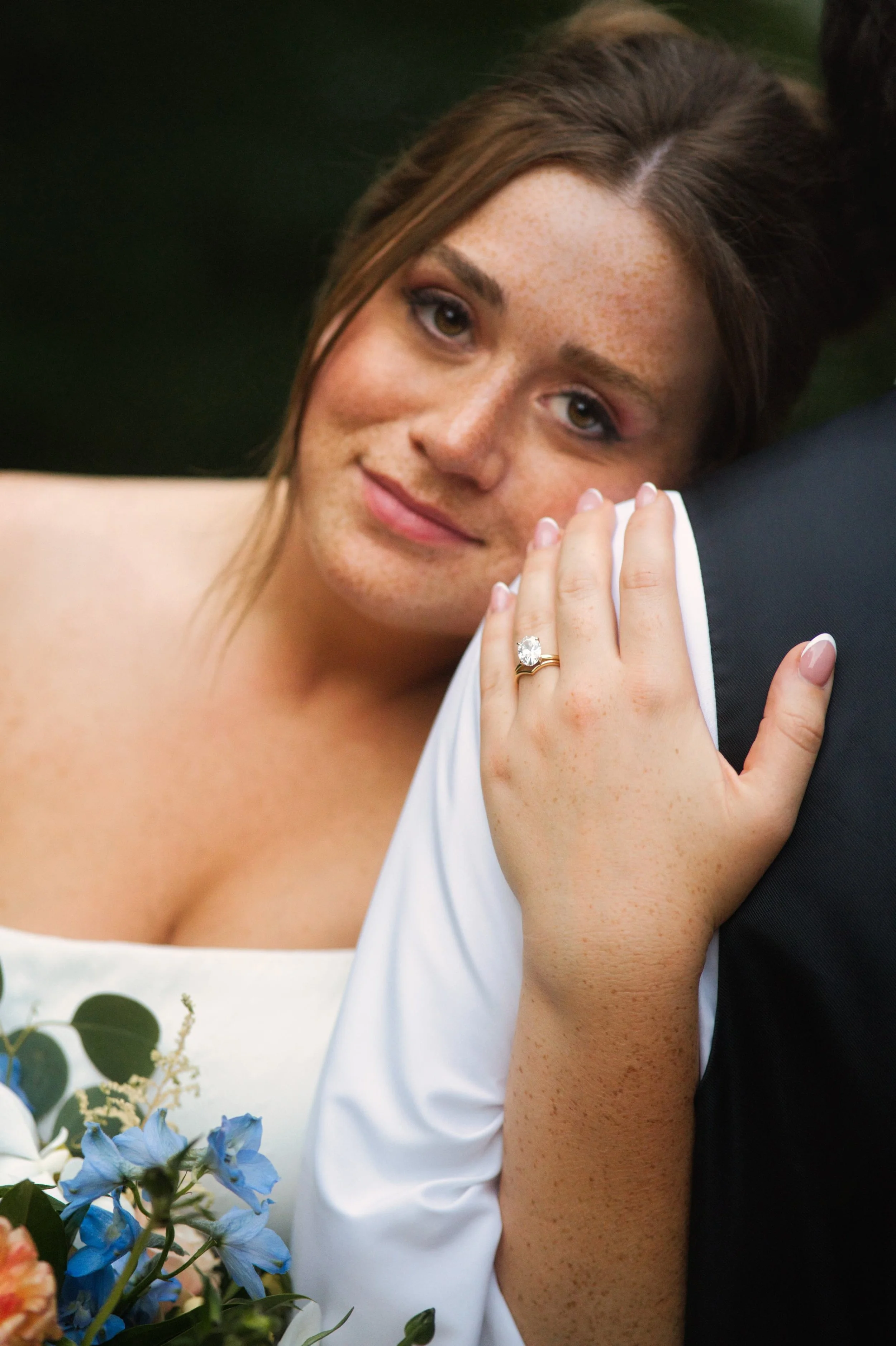 A woman with brown hair and freckles, smiling and resting her hand on a man's shoulder, showing an engagement ring.