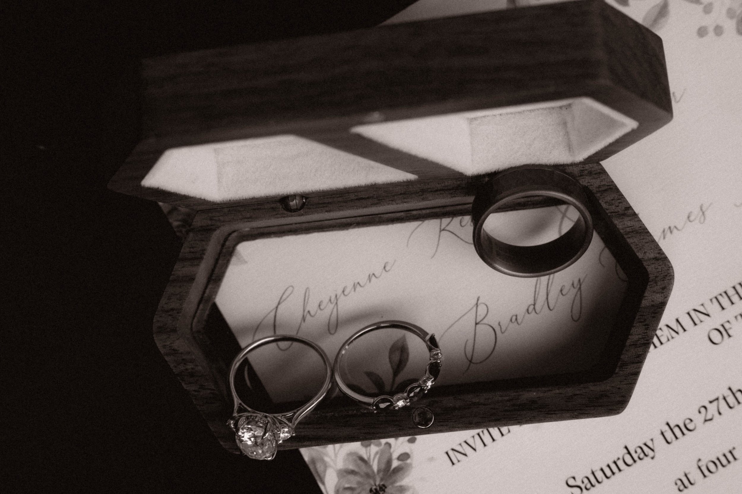 Round engagement ring with large center diamond and smaller side diamonds, and a wedding band with embedded diamonds, inside a wooden ring box with foam padding and a handwritten wedding invitation in the background.
