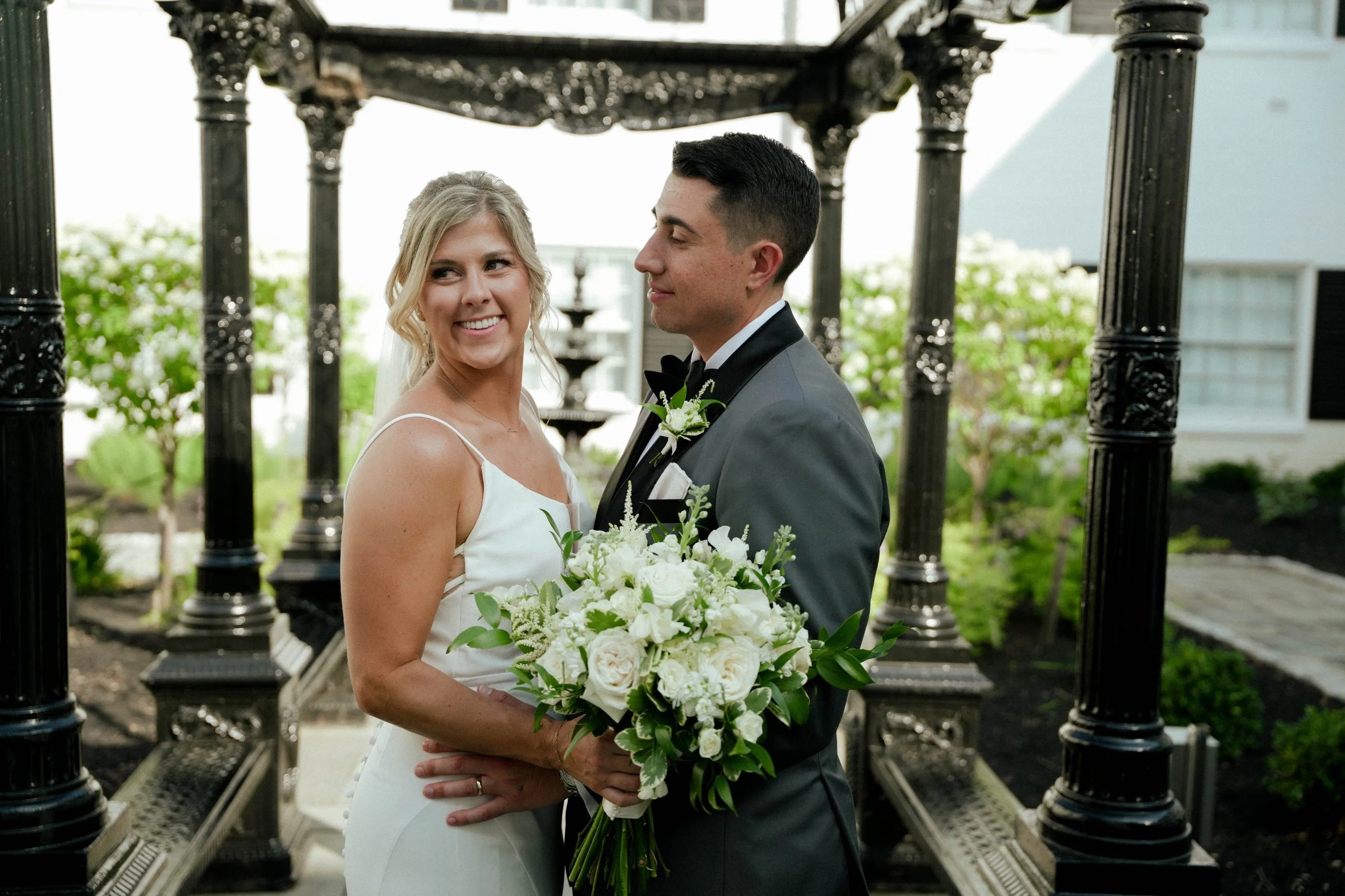 A bride and groom on their wedding day standing under a black ornate gazebo, with the bride holding a bouquet of white flowers and greenery, both smiling.